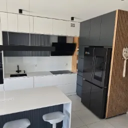 Nowoczesna kuchnia z białymi i grafitowymi szafkami, wyspą z białym blatem, czarną armaturą i lodówką side-by-side. Dekoracyjny panel drewniany z ozdobą w kształcie sowy.