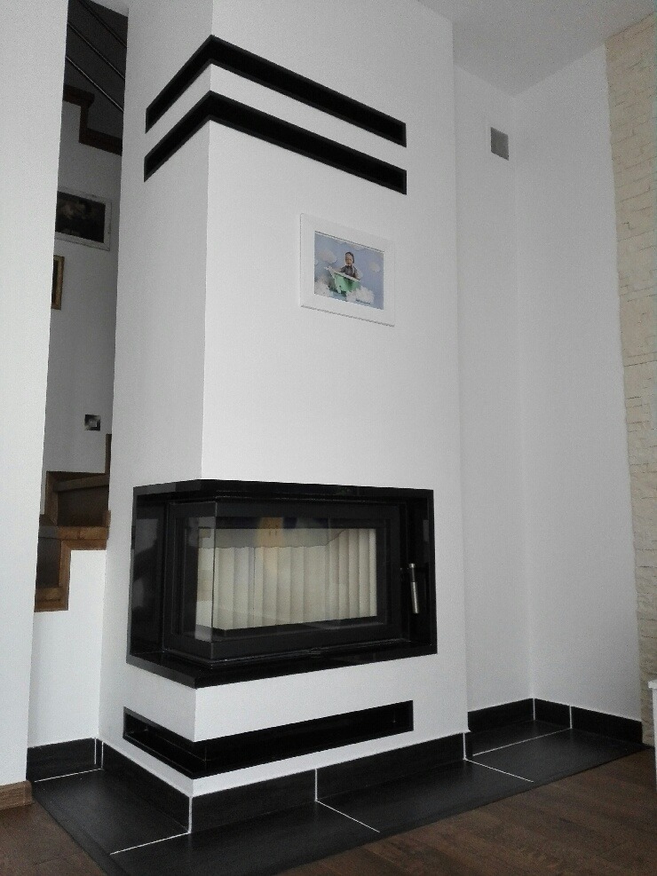 Biały kominek narożny z czarnymi akcentami, wbudowany w ścianę, z widocznym fragmentem schodów w tle.