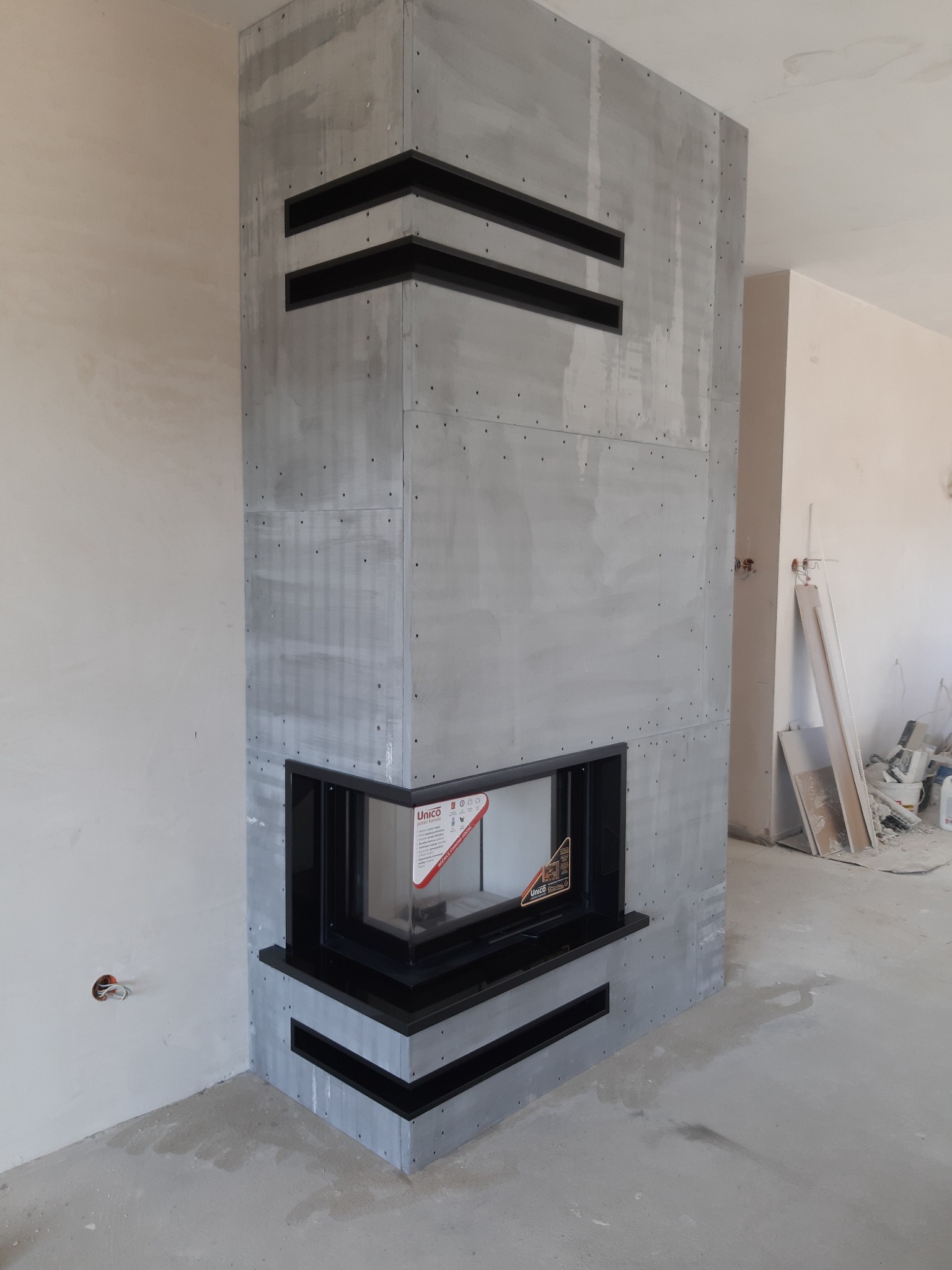 Wnętrze w trakcie wykańczania z widocznym obudowanym kominkiem o surowym, betonowym wykończeniu i czarnych akcentach, w tle widoczne materiały budowlane.