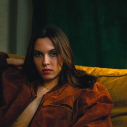 Emil Gawkowski - Portret kobiety w brązowej kurtce na musztardowej sofie przed zielonym tłem, z kolczykiem w nosie i delikatnym tatuażem na klatce piersiowej.