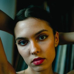 Emil Gawkowski - Portret młodej kobiety z piercingiem w nosie, patrzącej w obiektyw, z rękami uniesionymi za głową, w ciemnym otoczeniu.