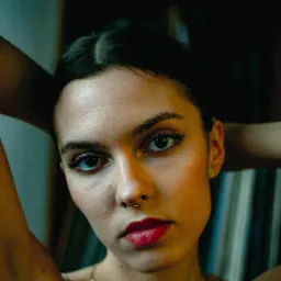 Portret młodej kobiety z piercingiem w nosie, patrzącej w obiektyw, z rękami uniesionymi za głową, w ciemnym otoczeniu.