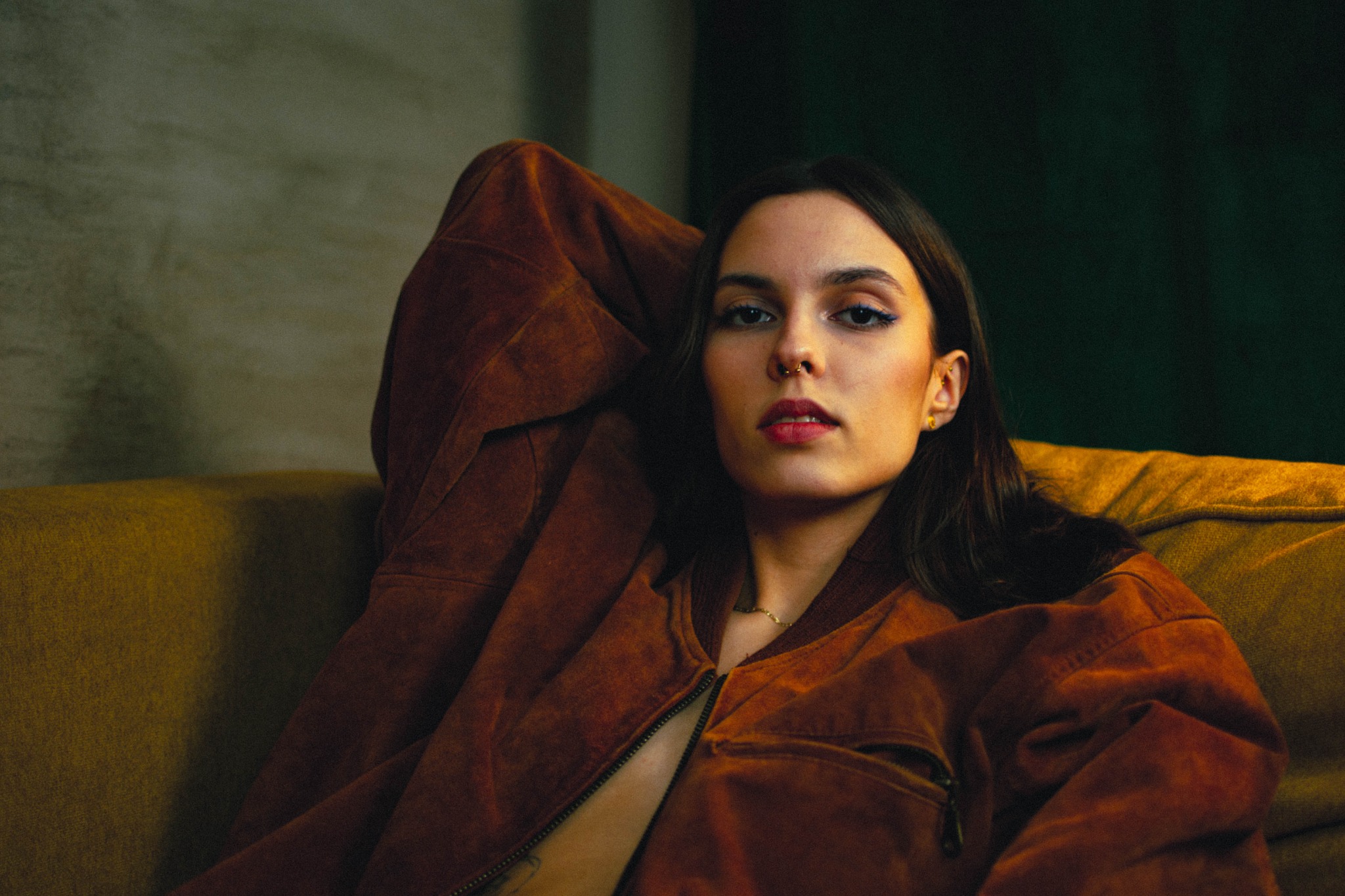 Portret kobiety w brązowej kurtce, z piercingiem w nosie, oparta o żółtą sofę na ciemnym tle.