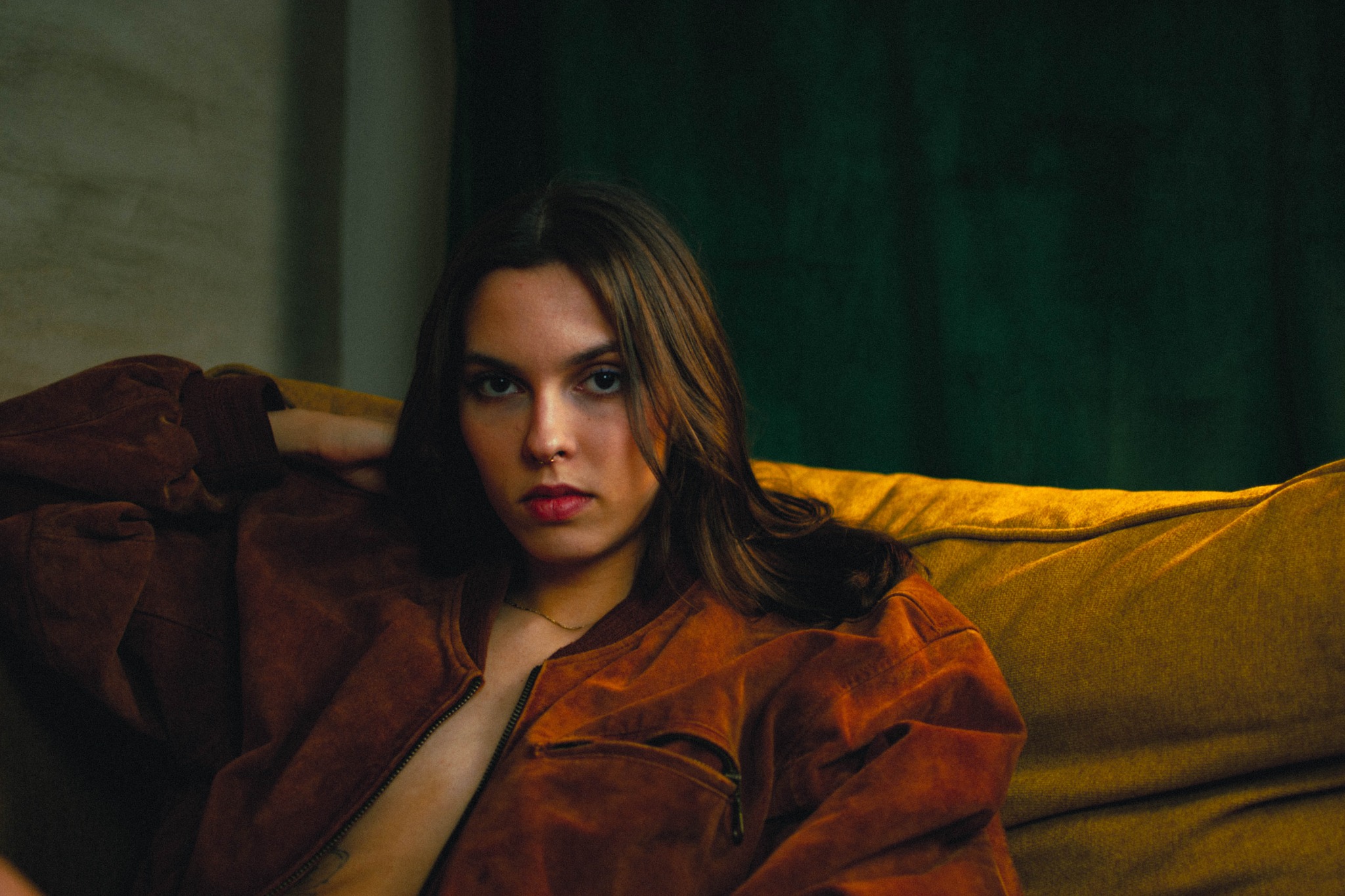 Portret kobiety w brązowej kurtce na musztardowej sofie przed zielonym tłem, z kolczykiem w nosie i delikatnym tatuażem na klatce piersiowej.