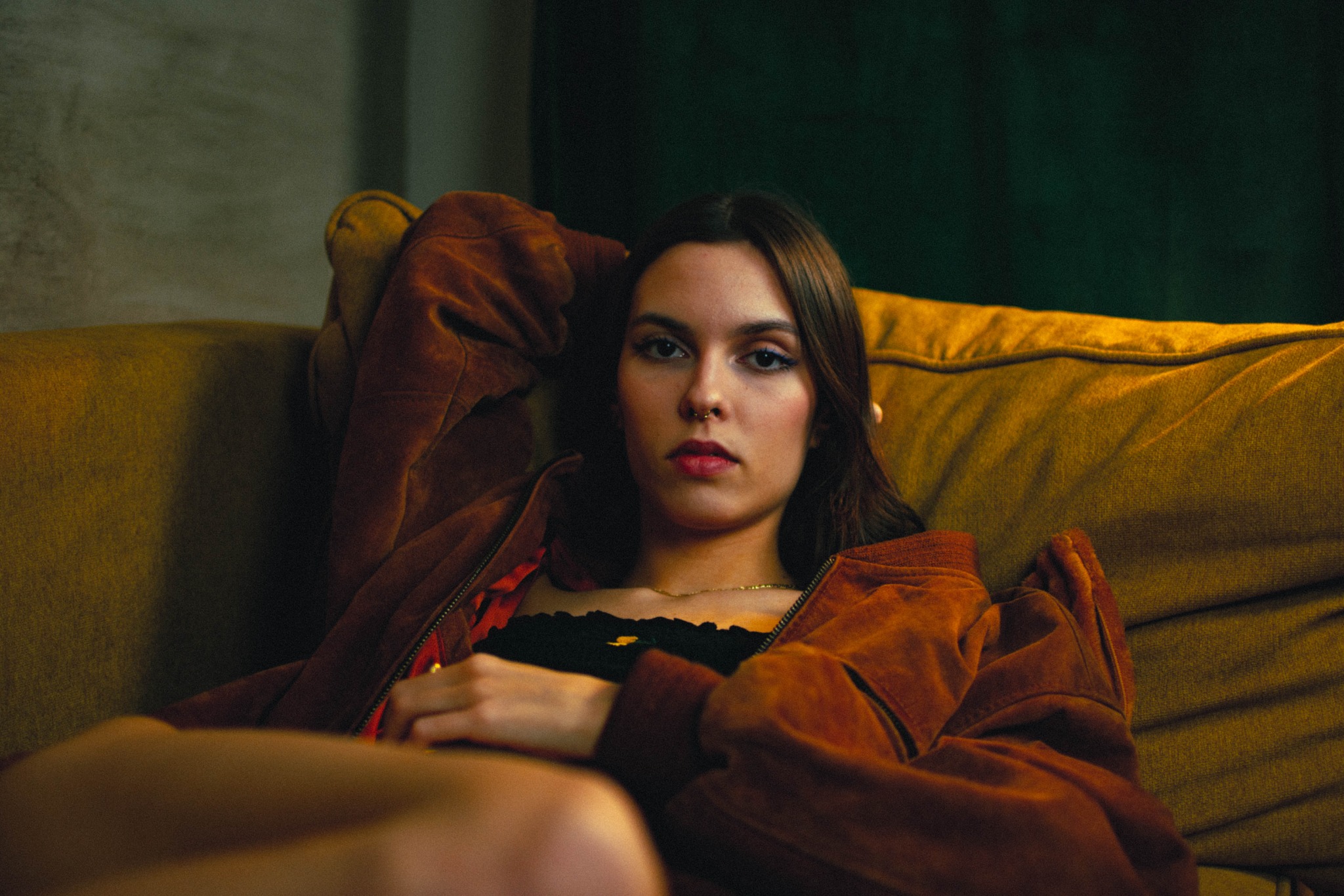Portret kobiety z piercingiem w nosie, w brązowej zamszowej kurtce, leżącej na musztardowej kanapie, z ciemnozielonym tłem.