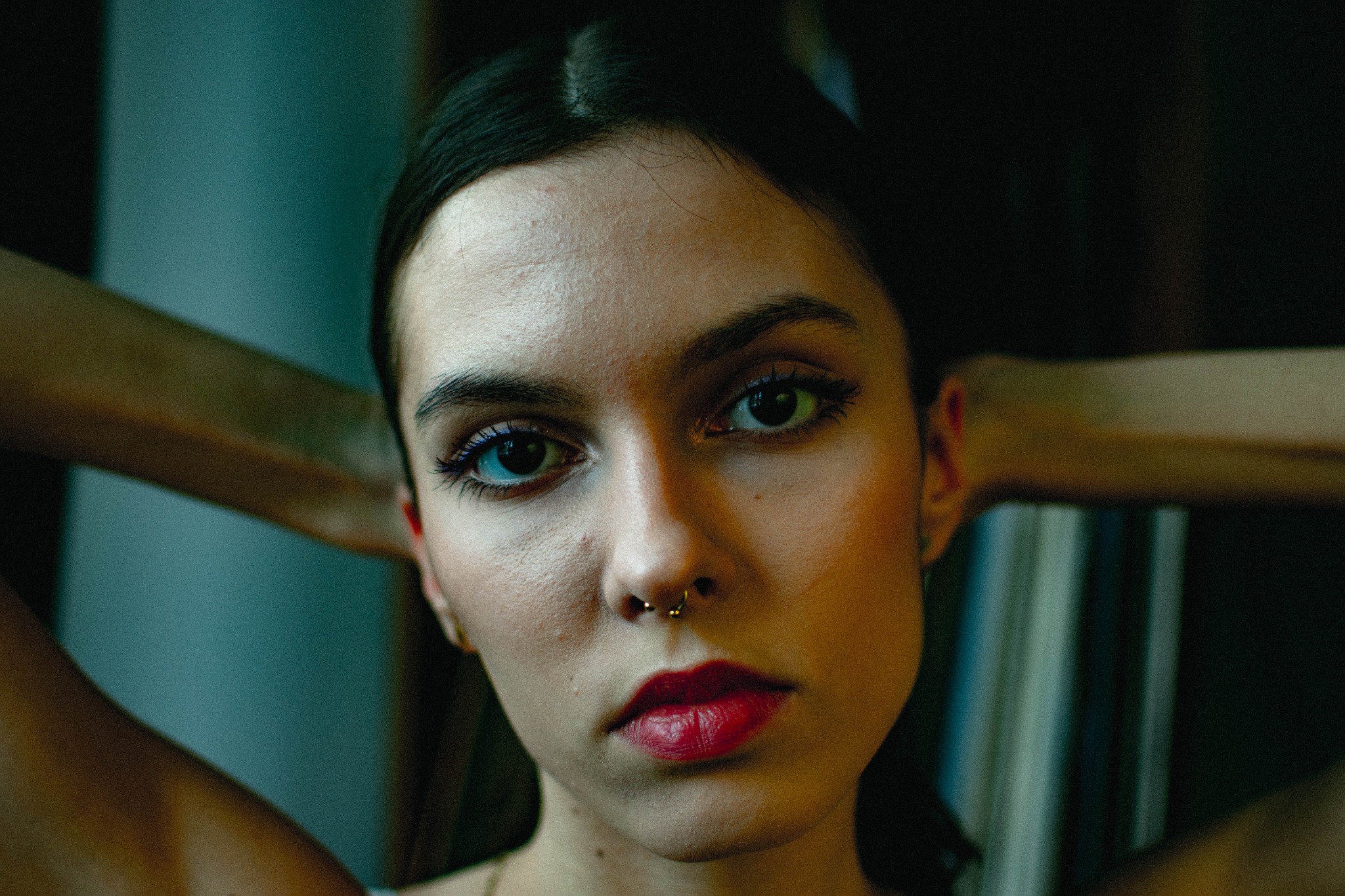 Portret młodej kobiety z piercingiem w nosie, intensywnym makijażem oczu i czerwonymi ustami, ręce uniesione za głową, ciemne tło.