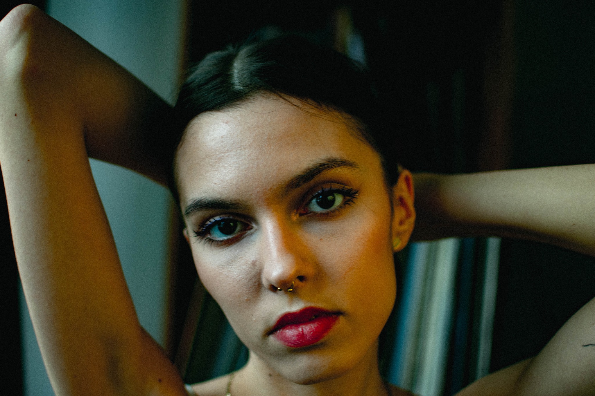 Portret młodej kobiety z piercingiem w nosie, patrzącej w obiektyw, z rękami uniesionymi za głową, w ciemnym otoczeniu.