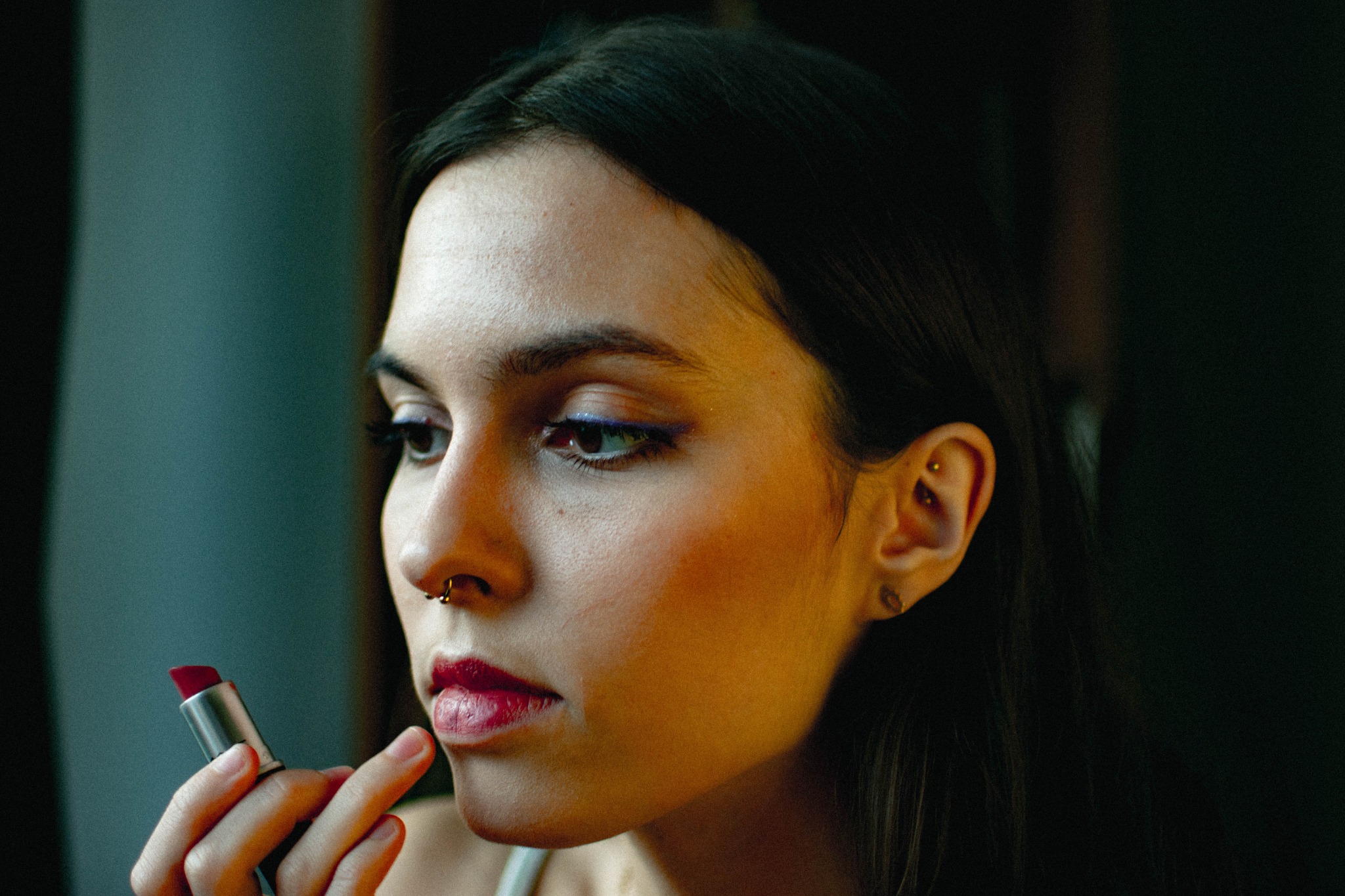 Portret kobiety z piercingiem w nosie i uchu, nakładającej czerwoną szminkę, z widoczną niebieską kreską na powiece.