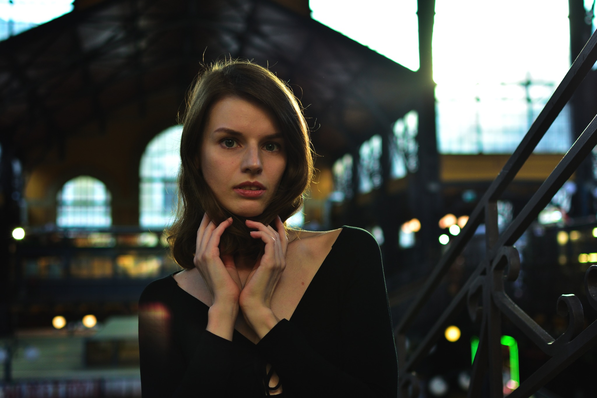 Portret młodej kobiety w czarnej sukience na tle zabytkowej architektury z oknami łukowymi i metalową balustradą, oświetlonej ciepłym światłem.