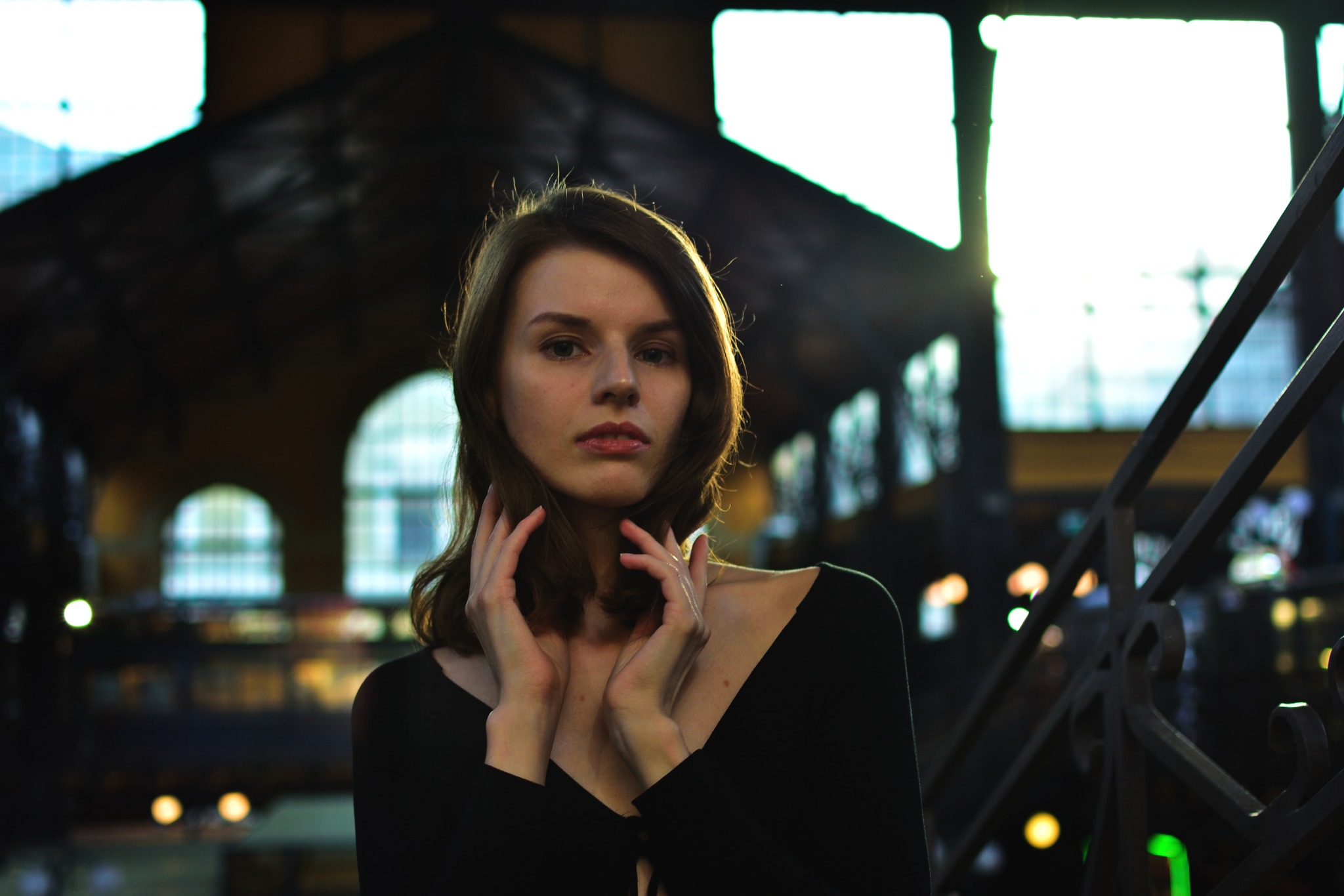 Portret młodej kobiety w czarnej bluzce na tle hali targowej z oknami łukowymi i metalową balustradą