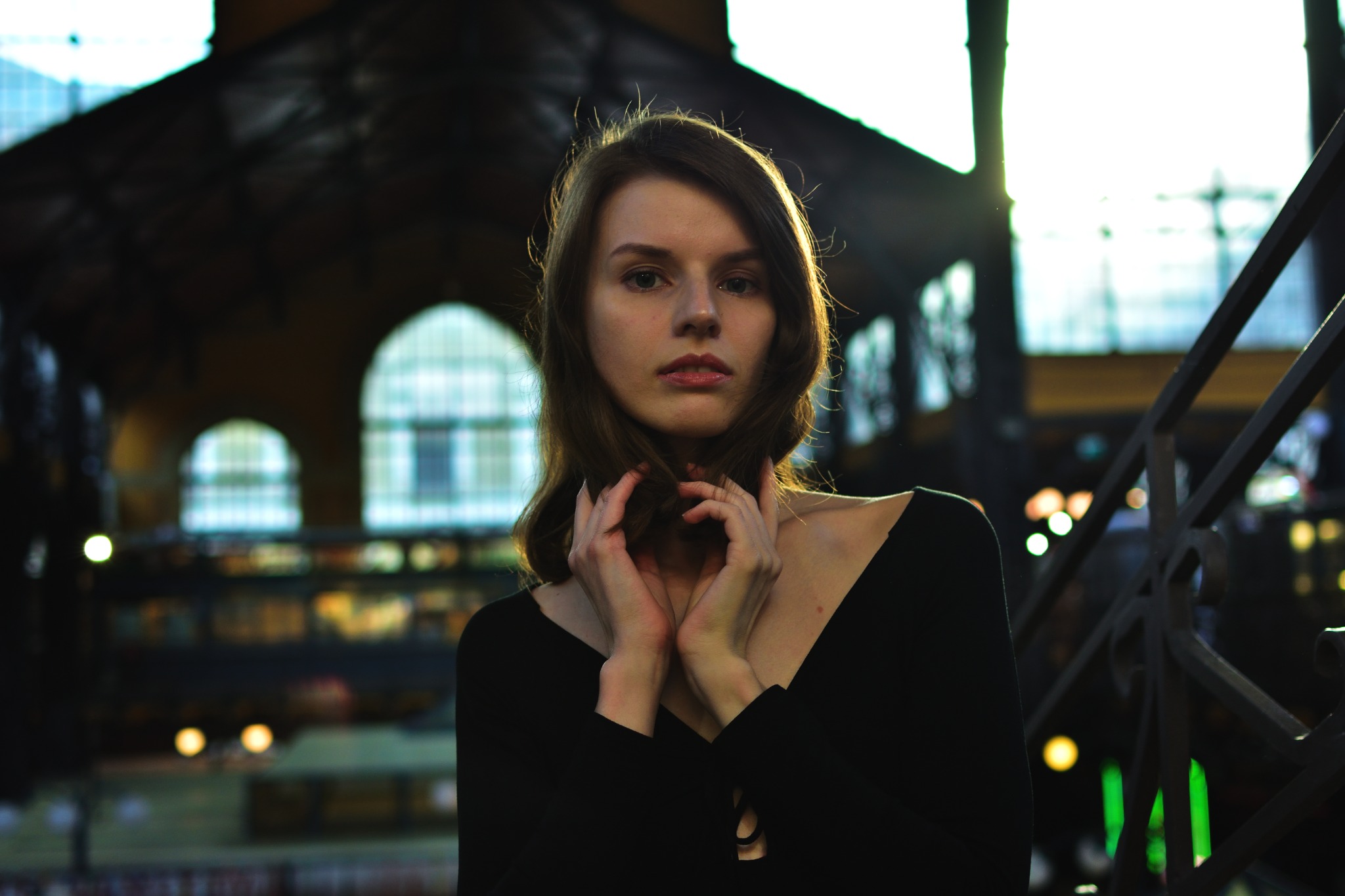 Portret młodej kobiety w czarnej sukience na tle hali z oknami łukowymi, dłonie uniesione do włosów, delikatne światło.