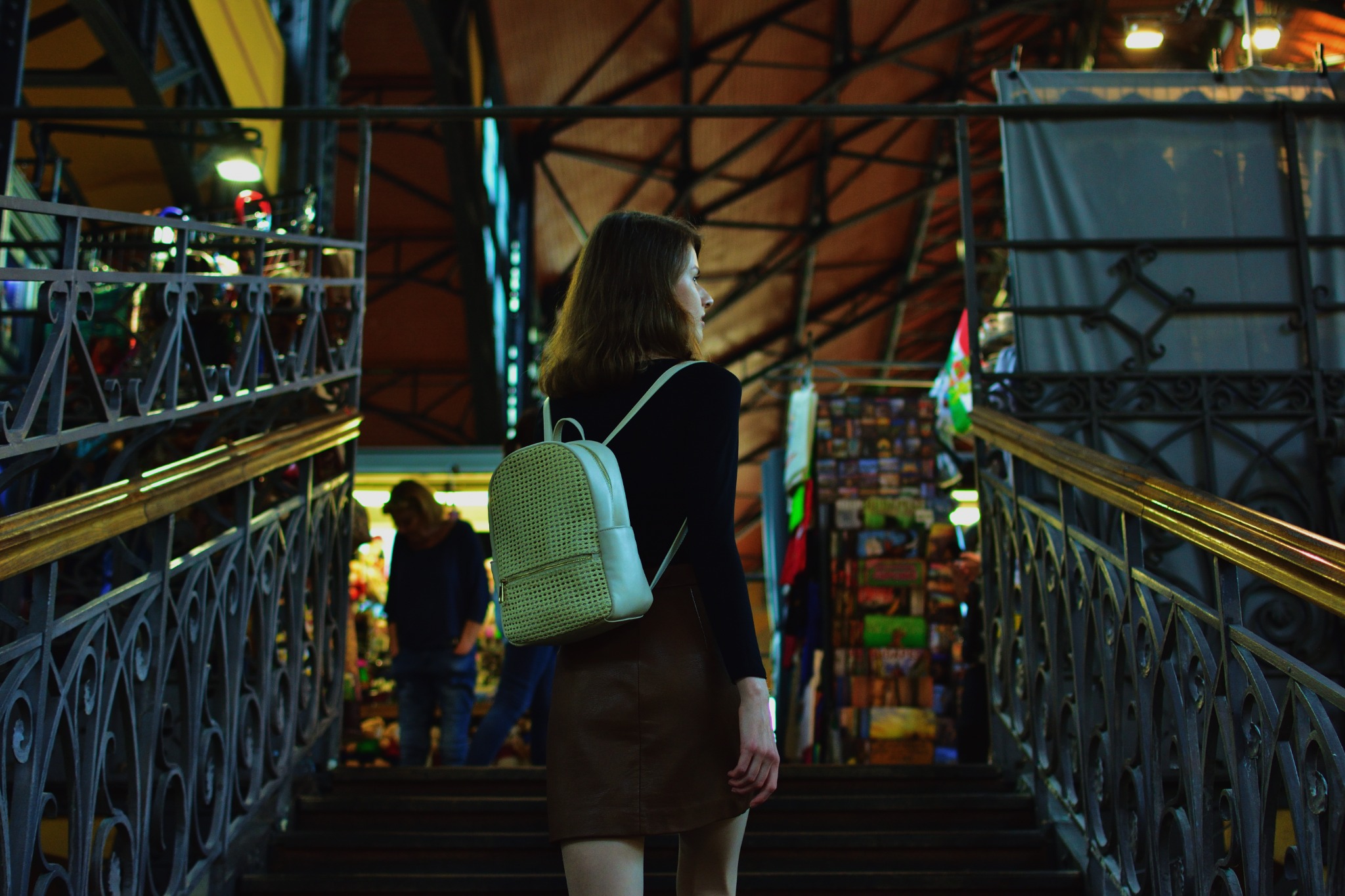 Kobieta z plecakiem w brązowej spódnicy wchodzi po schodach z metalową balustradą w hali targowej z widoczną konstrukcją dachu i stoiskami w tle.