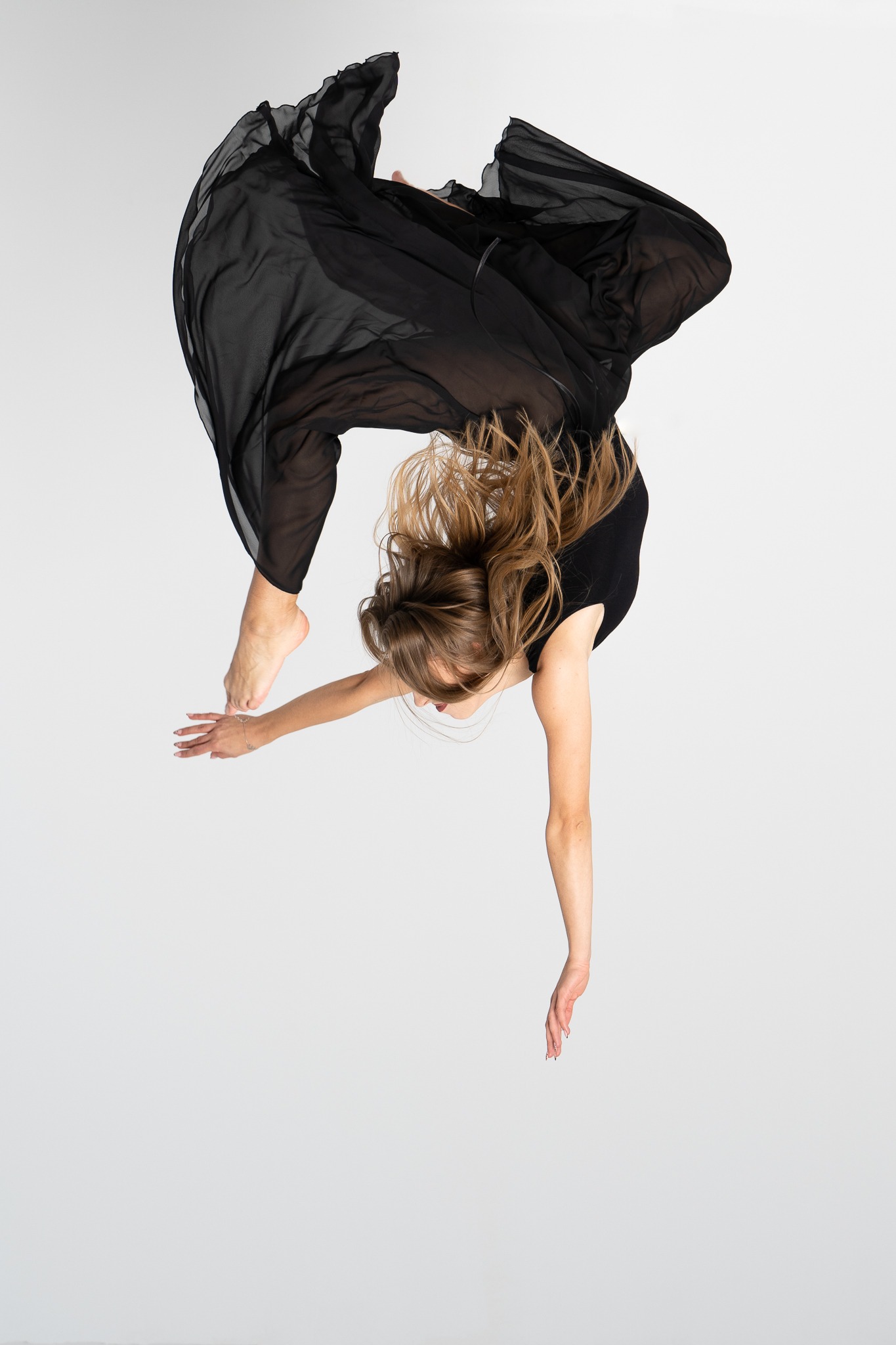 Kobieta w czarnym stroju i zwiewnej sukience, w dynamicznej pozie, unosząca się w powietrzu na jasnym tle, z widocznym drobnym srebrnym łańcuszkiem na nadgarstku.