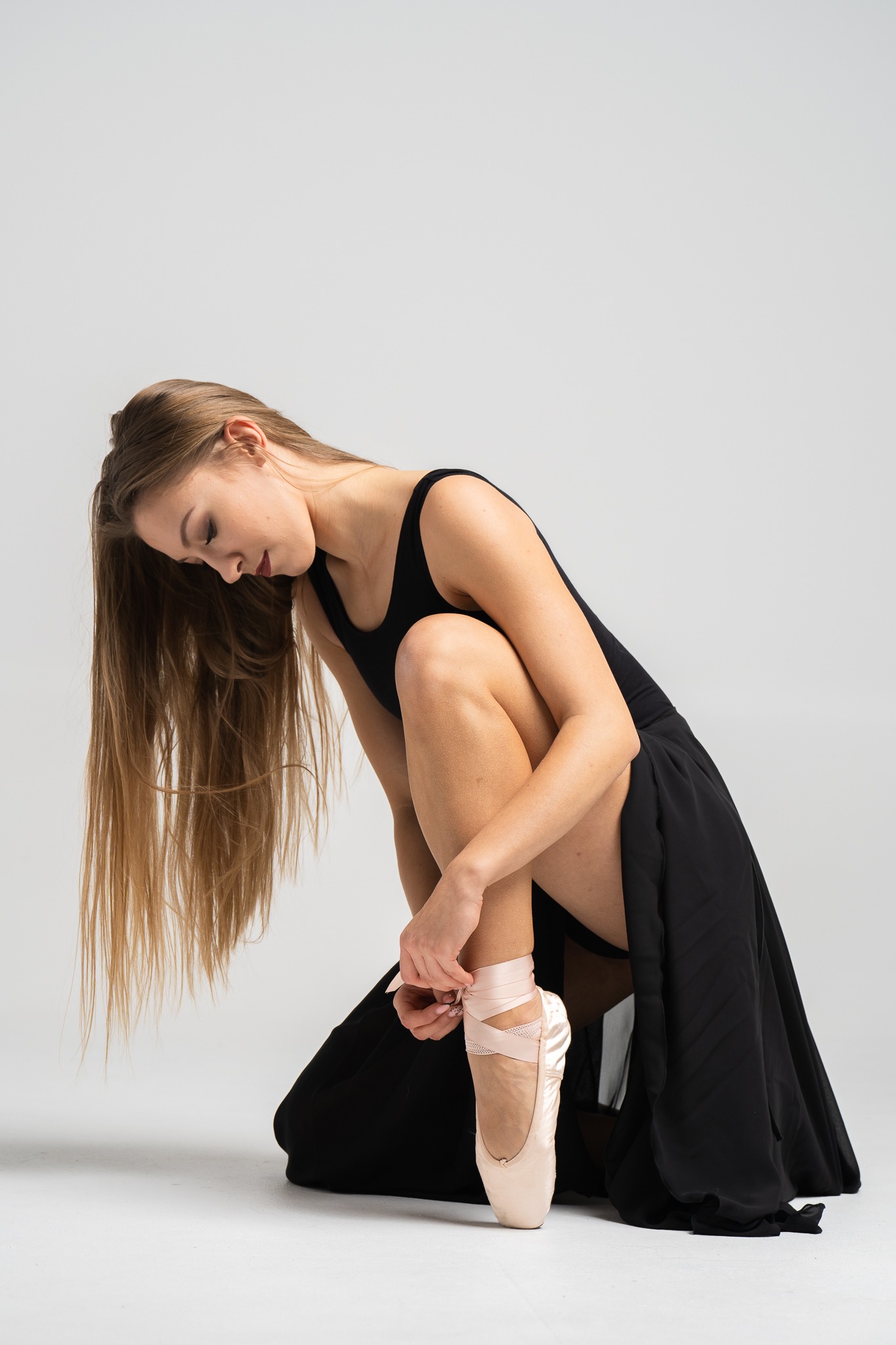 Baletnica wiąże wstążki pointów, pochylona w czarnym stroju na jasnym tle, z długimi blond włosami opadającymi na bok.