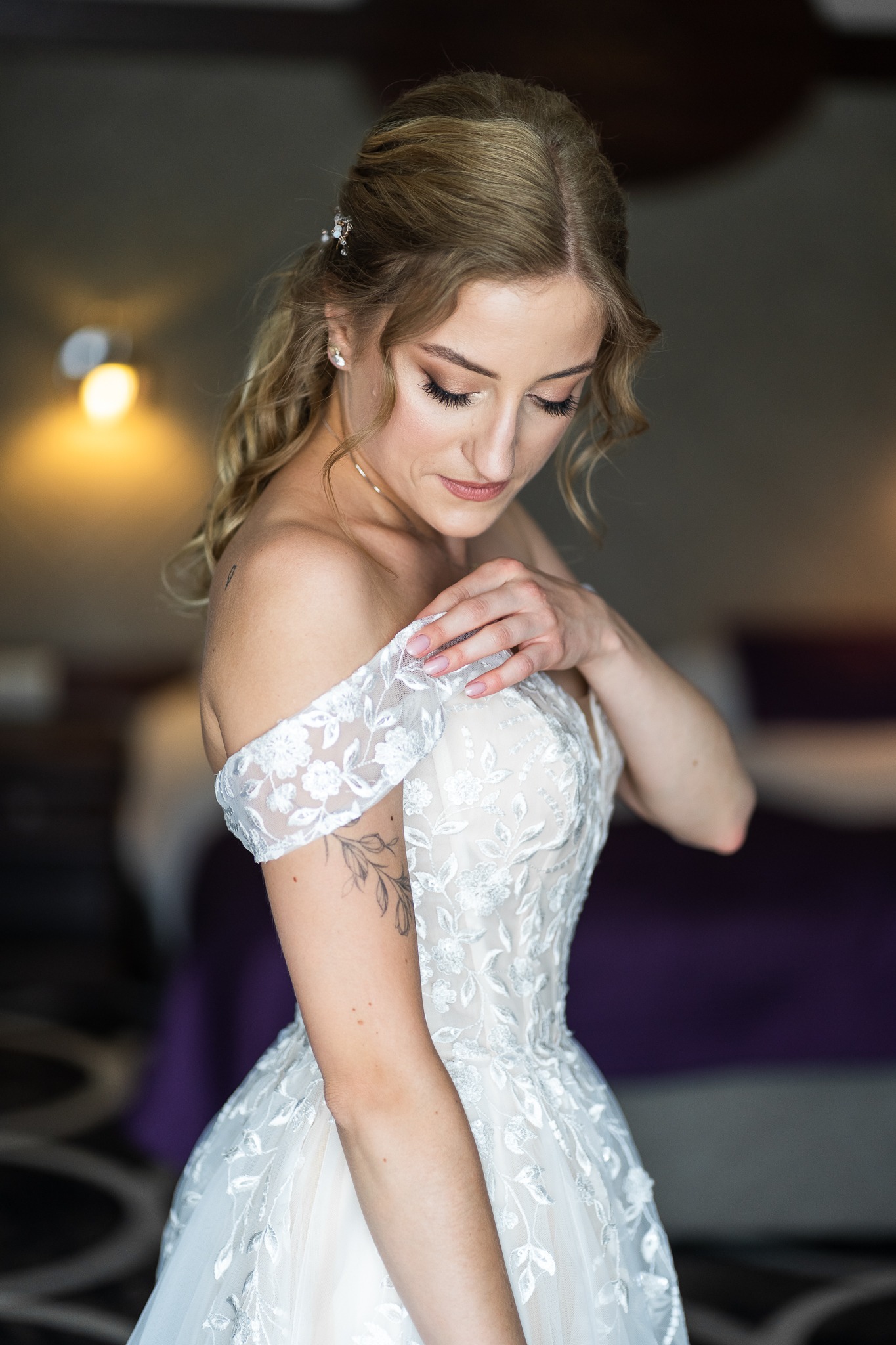Panna młoda w białej sukni ślubnej z odkrytymi ramionami, dotykająca delikatnie dekoltu, z widocznym tatuażem na ramieniu i upiętymi blond włosami z ozdobną spinką.