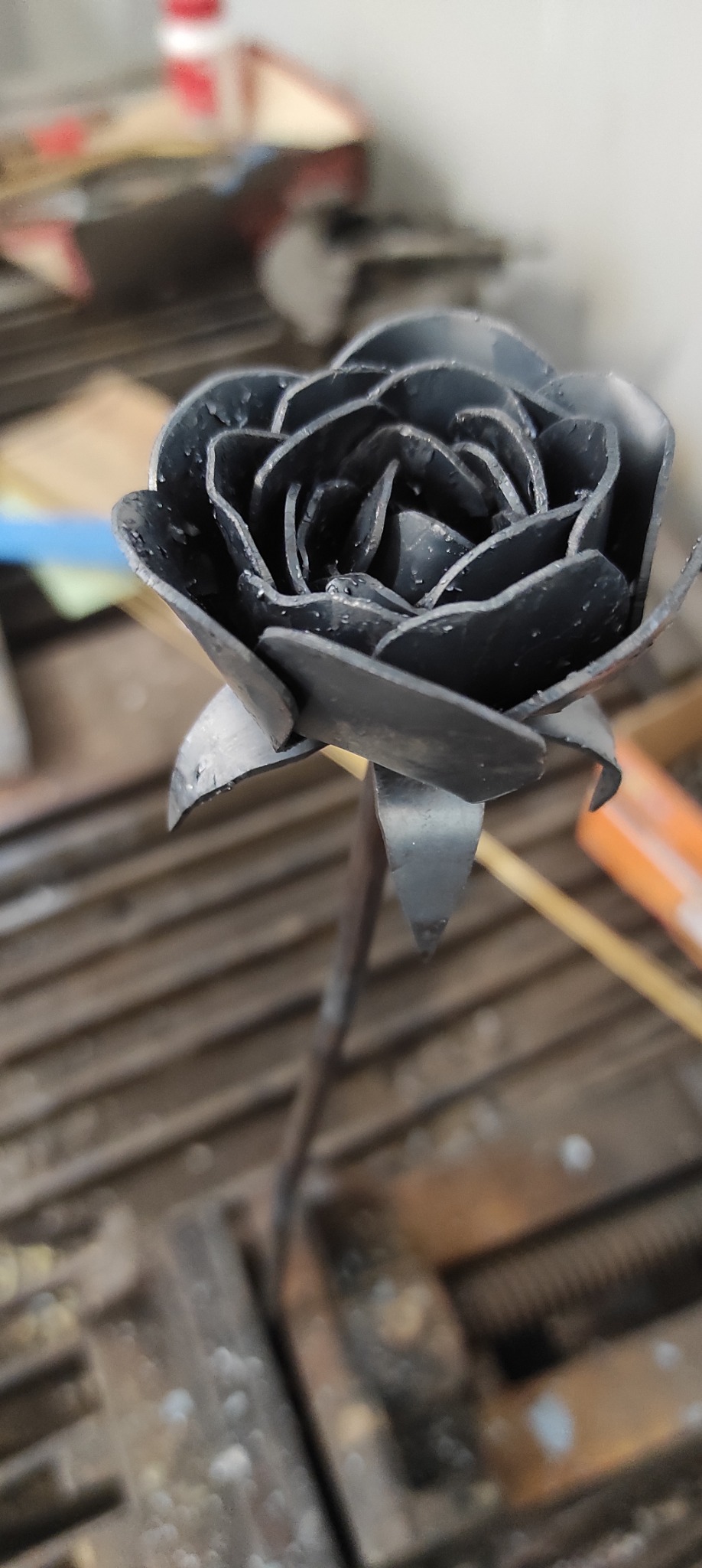 Ręcznie wykonana metalowa róża z widocznymi śladami spawania na płatkach, umieszczona na warsztatowym stole z metalowymi szczeblami.