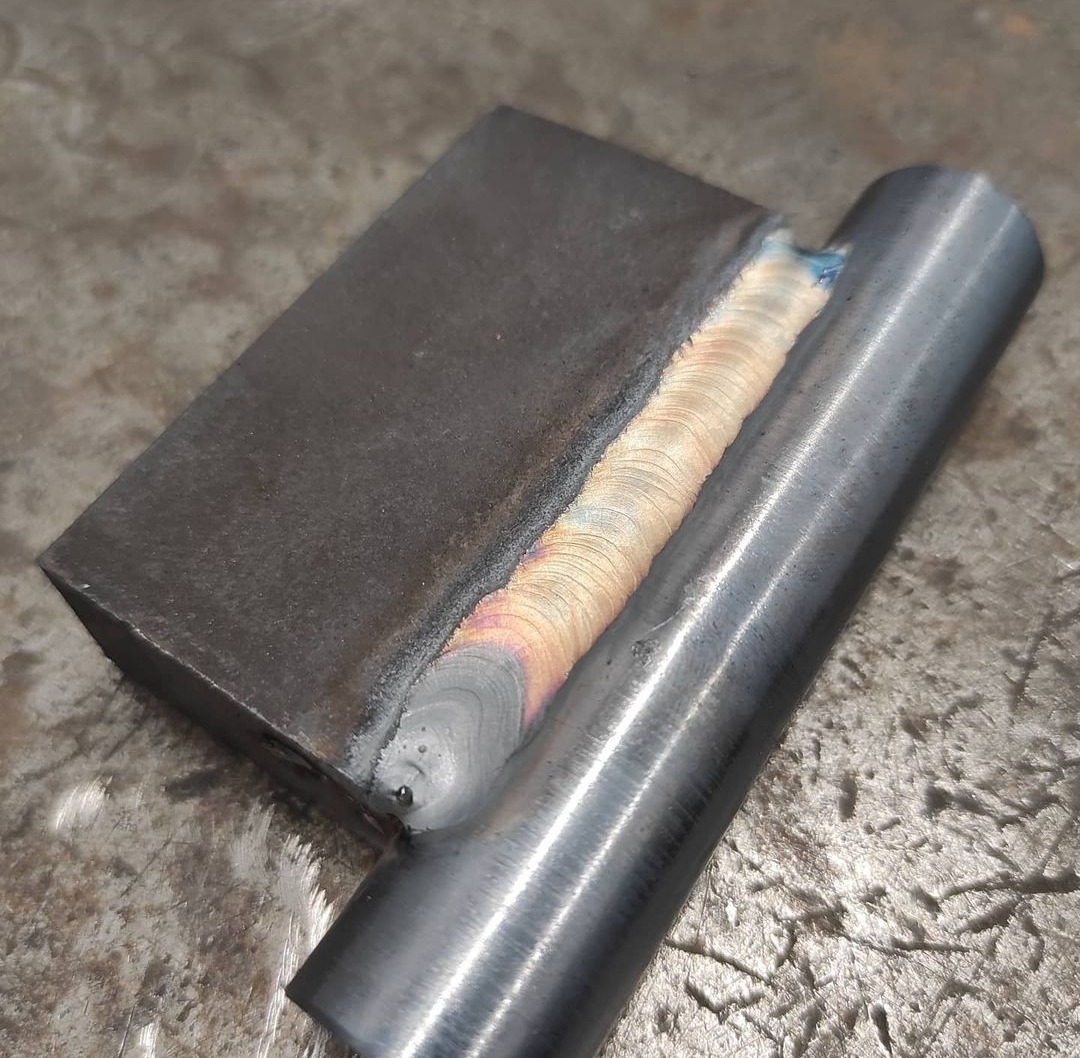 Połączenie spawane blachy i rury stalowej, widoczna precyzyjna spoina z opalizującymi przebarwieniami termicznymi.