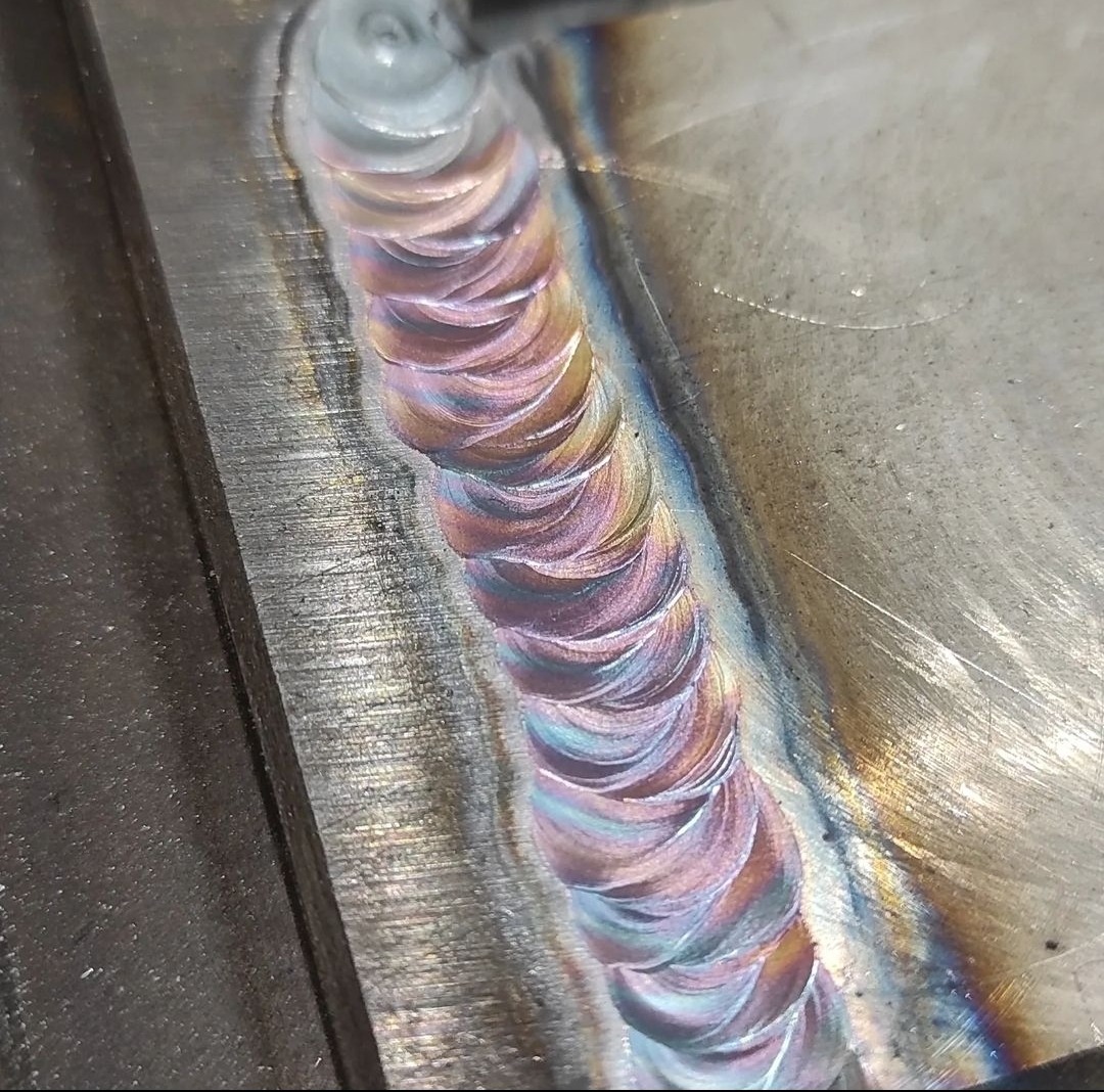 Świeżo wykonany spaw na metalowej powierzchni, z widocznymi tęczowymi przebarwieniami termicznymi i charakterystycznym wzorem nakładających się półksiężyców.