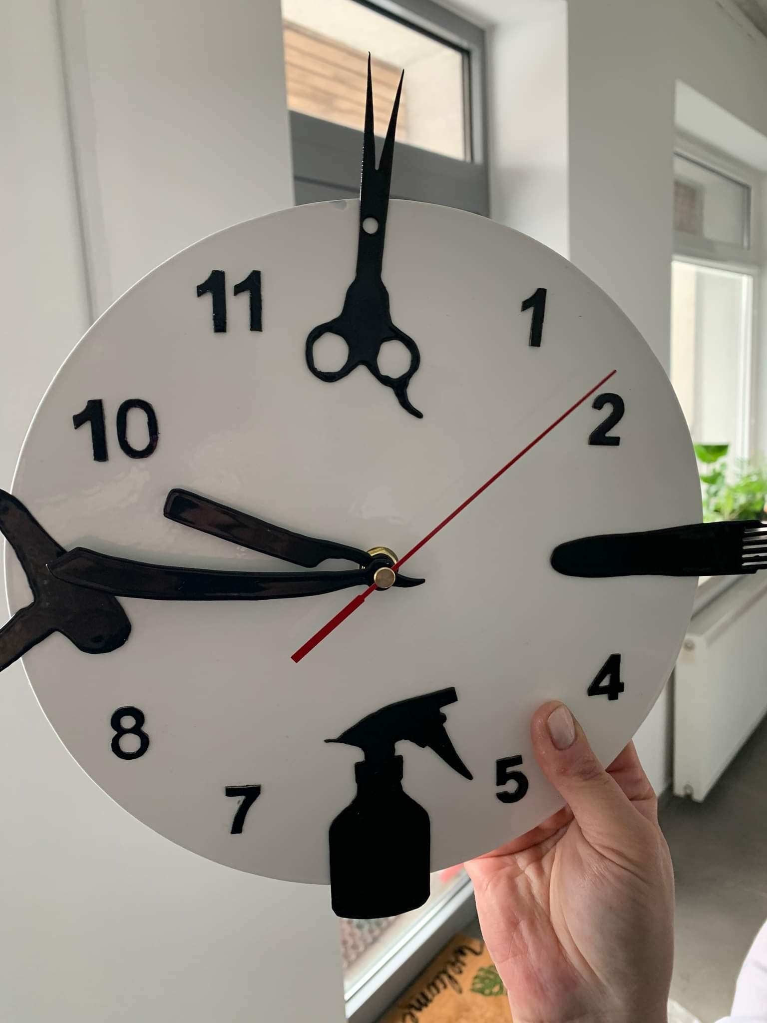 Okrągły zegar z czarnymi symbolami fryzjerskimi zamiast cyfr: nożyczki, brzytwa, grzebień i spryskiwacz, trzymany w dłoni na tle jasnego wnętrza.