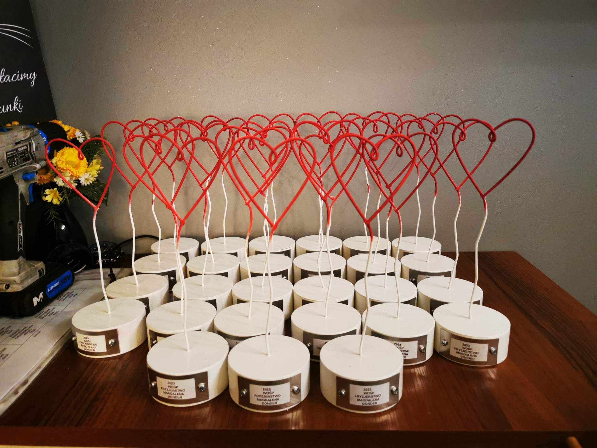 Ręcznie robione, metalowe serca na białych podstawkach z przymocowanymi tabliczkami, ustawione rzędami na drewnianym stole, obok wiertarko-wkrętarki i kwiatów.