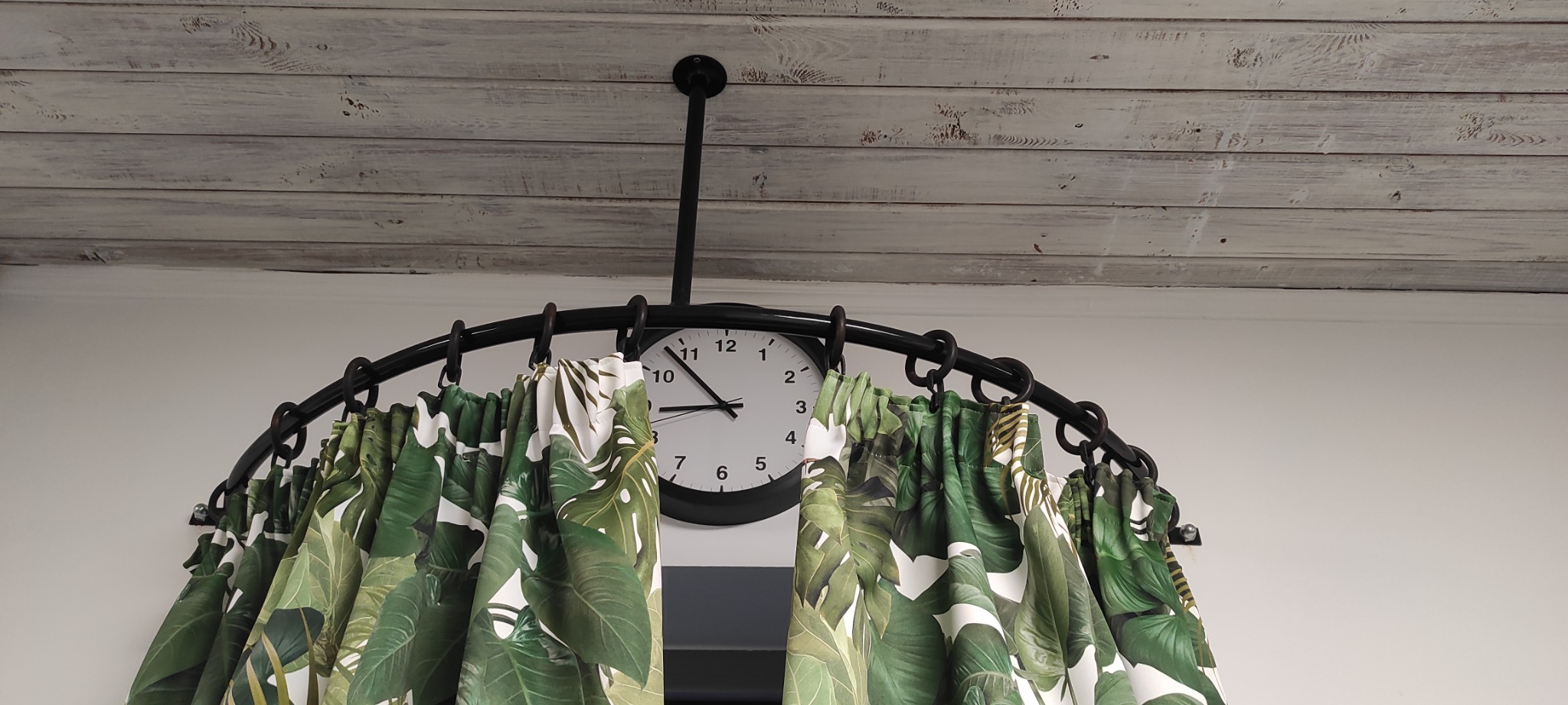 Czarny, metalowy karnisz łukowy z zegarem w centralnej części, zawieszony na tle białej, drewnianej boazerii i zielonych zasłon w motywy roślinne.