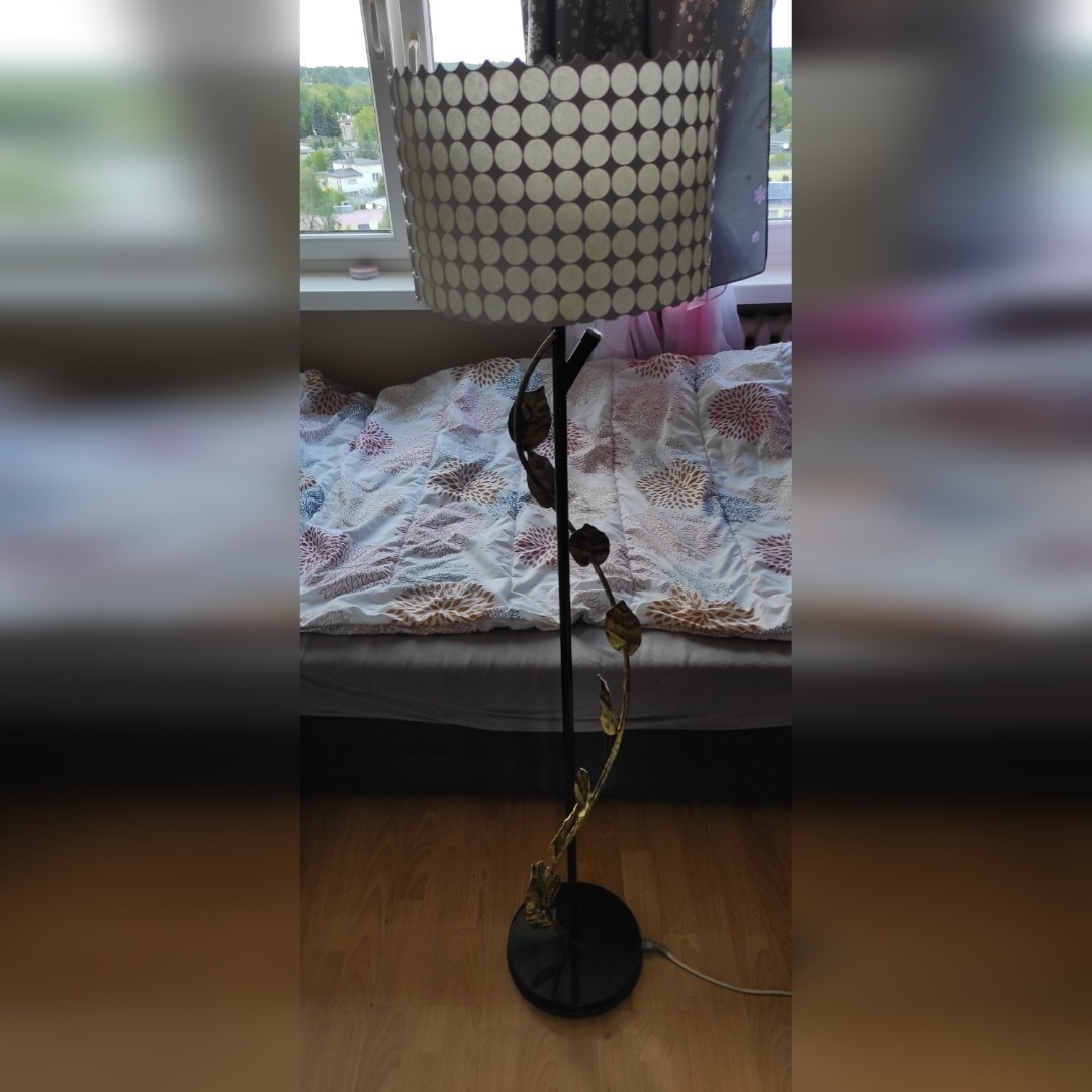 Stojąca lampa podłogowa z abażurem w kropki i metalową konstrukcją imitującą roślinę z liśćmi, ustawiona na drewnianej podłodze w pokoju dziennym z widokiem na zewnątrz przez okno.