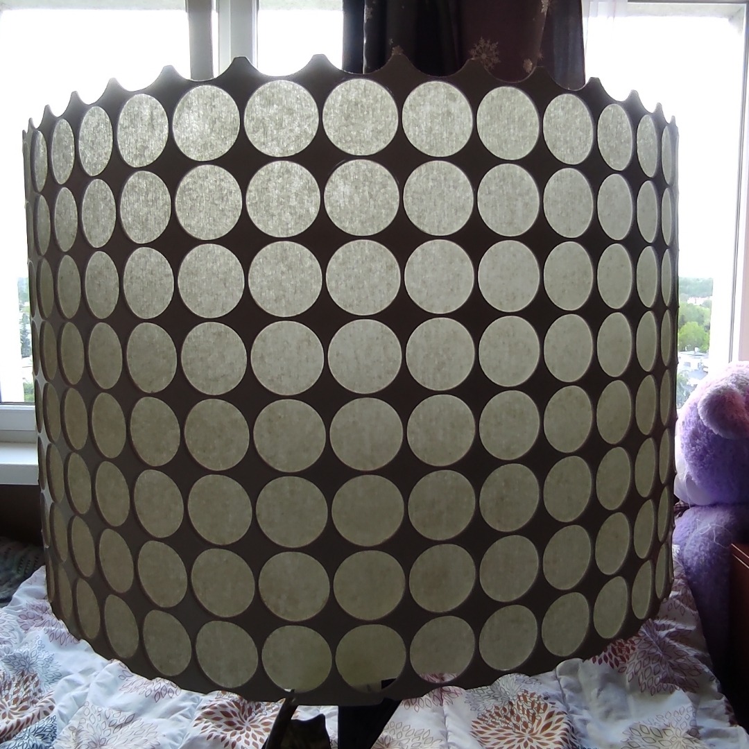 Abażur lampy z wyciętymi otworami w kształcie kół, stojący na wzorzystej pościeli, z widocznym oknem w tle.