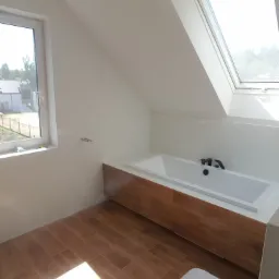 Jasna łazienka na poddaszu z drewnopodobną zabudową wanny, brązowymi płytkami podłogowymi i oknem dachowym, widok na częściowo zamontowaną toaletę.