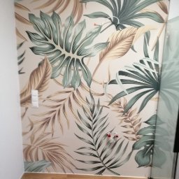 Adbud - Ściana z fototapetą w tropikalne liście, widoczne gniazdka elektryczne i fragment kabiny prysznicowej, panele podłogowe.