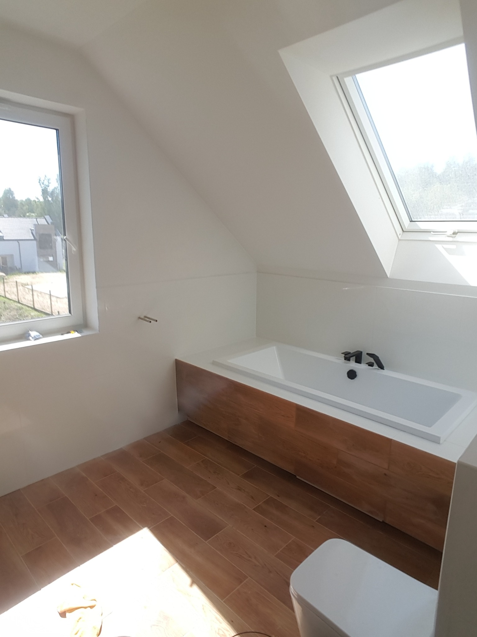 Jasna łazienka na poddaszu z drewnopodobną zabudową wanny, brązowymi płytkami podłogowymi i oknem dachowym, widok na częściowo zamontowaną toaletę.