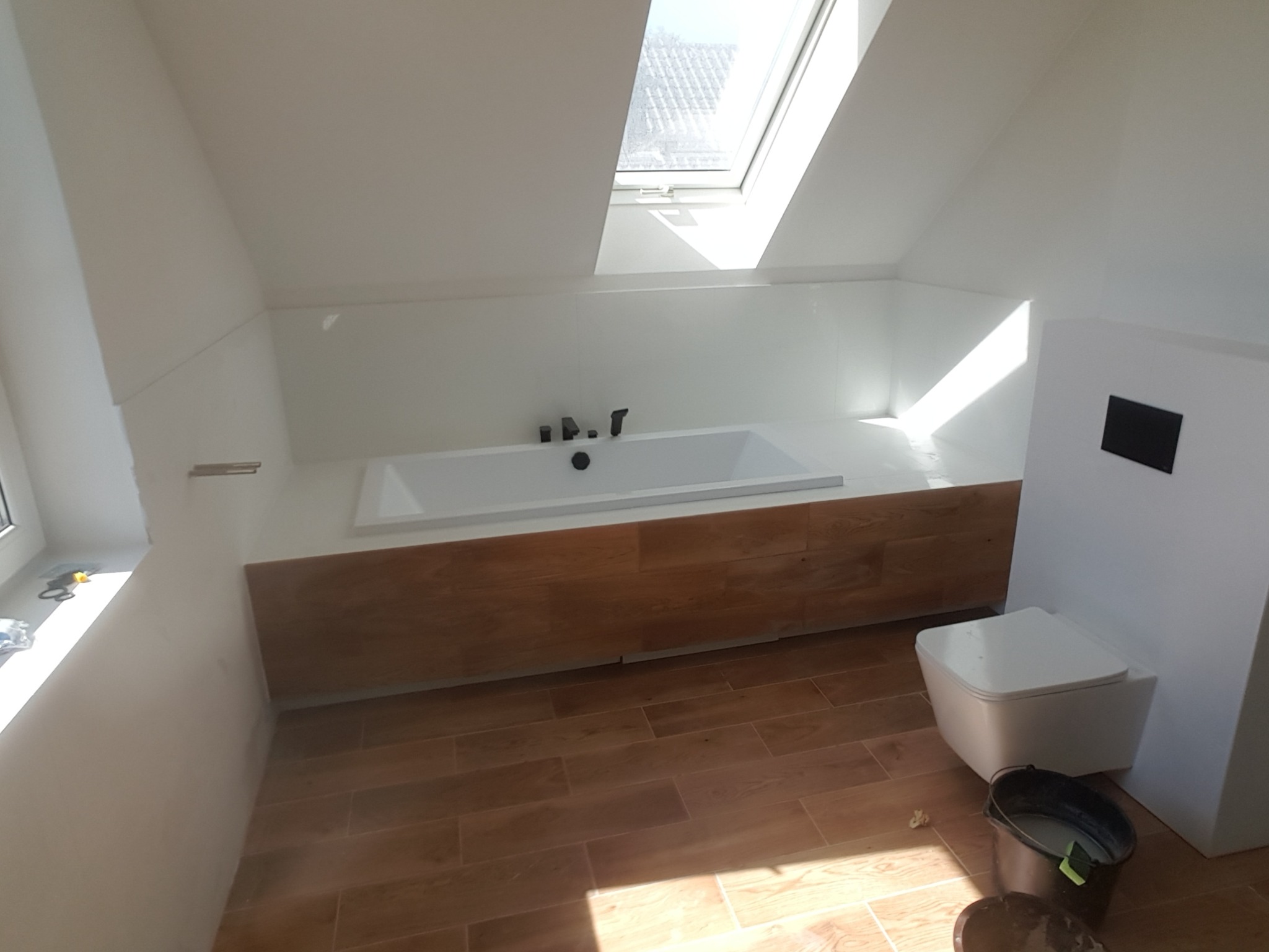 Nowoczesna łazienka na poddaszu z oknem dachowym, biała wanna obudowana drewnem, podwieszana toaleta, drewnopodobna podłoga, wiadro z zaprawą w rogu.