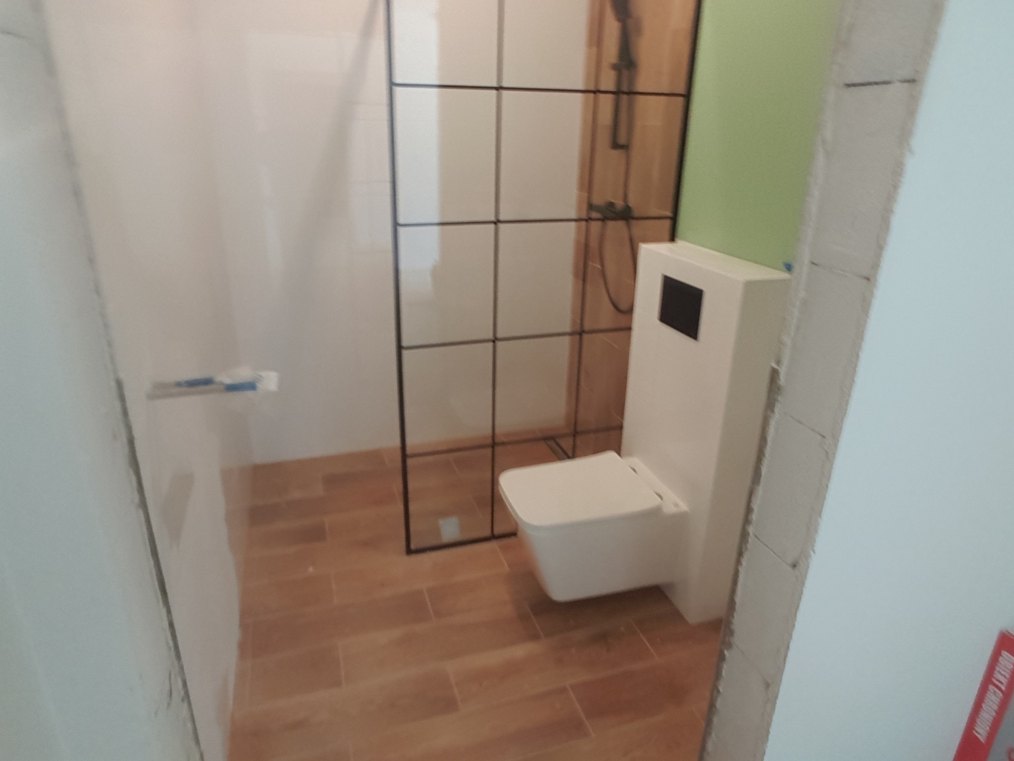 Nowoczesna łazienka w trakcie wykończenia: podwieszana toaleta, kabina prysznicowa z czarnymi profilami, drewnopodobna terakota na podłodze, ściany częściowo przygotowane do malowania.