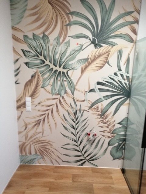 Ściana z fototapetą w tropikalne liście, widoczne gniazdka elektryczne i fragment kabiny prysznicowej, panele podłogowe.