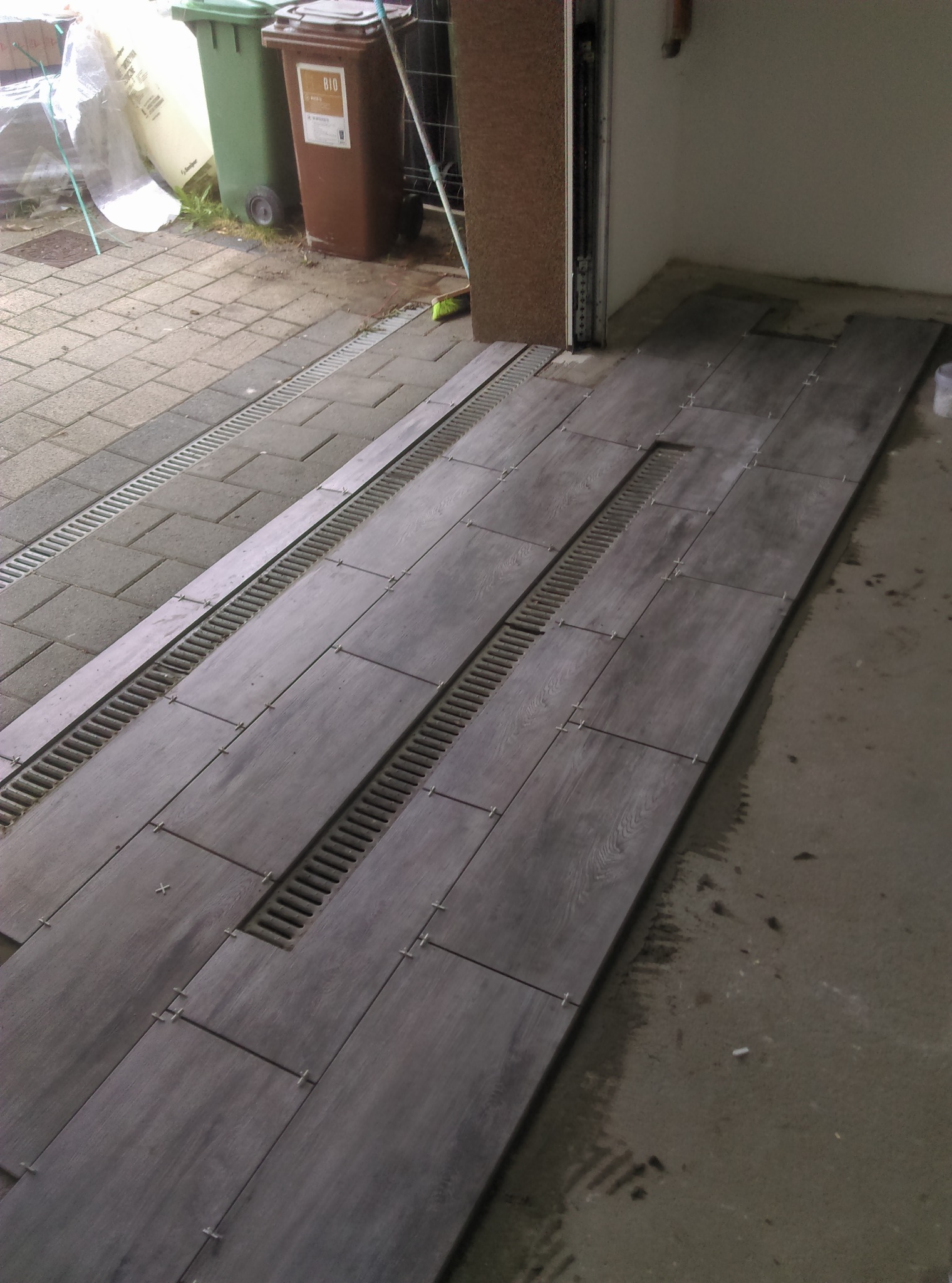 Montaż podłogi w garażu: ciemnoszare, prostokątne płytki imitujące drewno z krzyżykami dystansowymi, ułożone wzdłuż kanałów odwadniających. Część płytek jeszcze nieułożona, widoczna surowa posadzka...