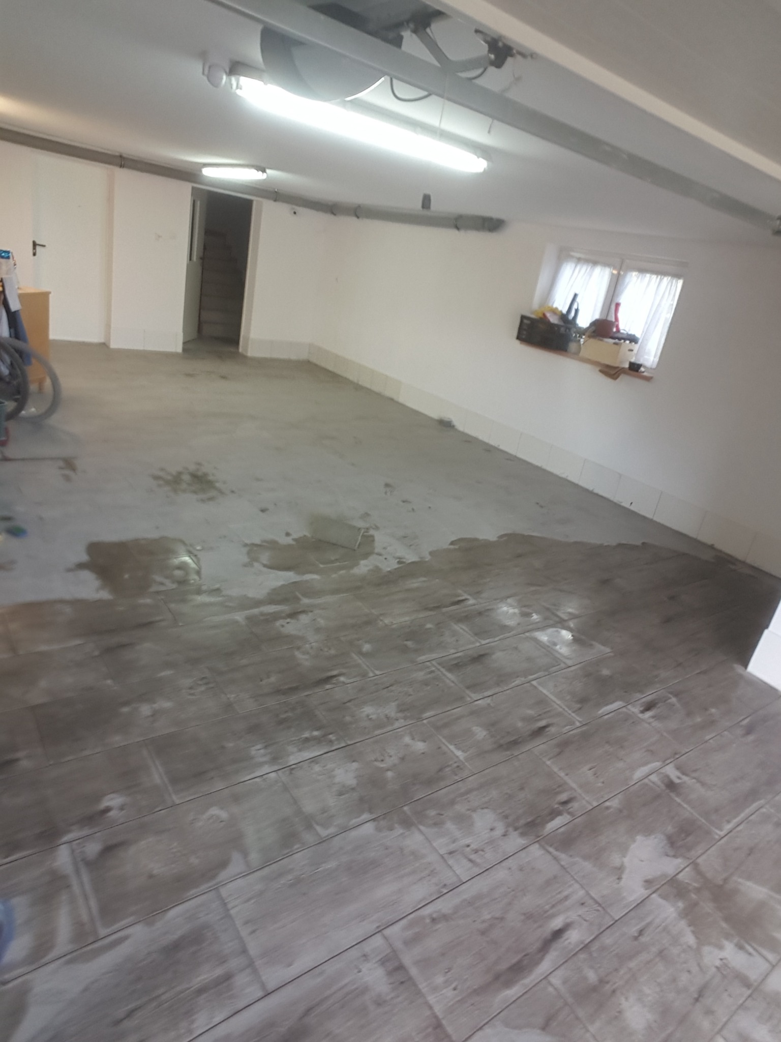 Układanie płytek podłogowych imitujących drewno w surowym stanie, z widocznymi plamami po kleju lub zaprawie, w pomieszczeniu z białymi ścianami i oknem z firanką.