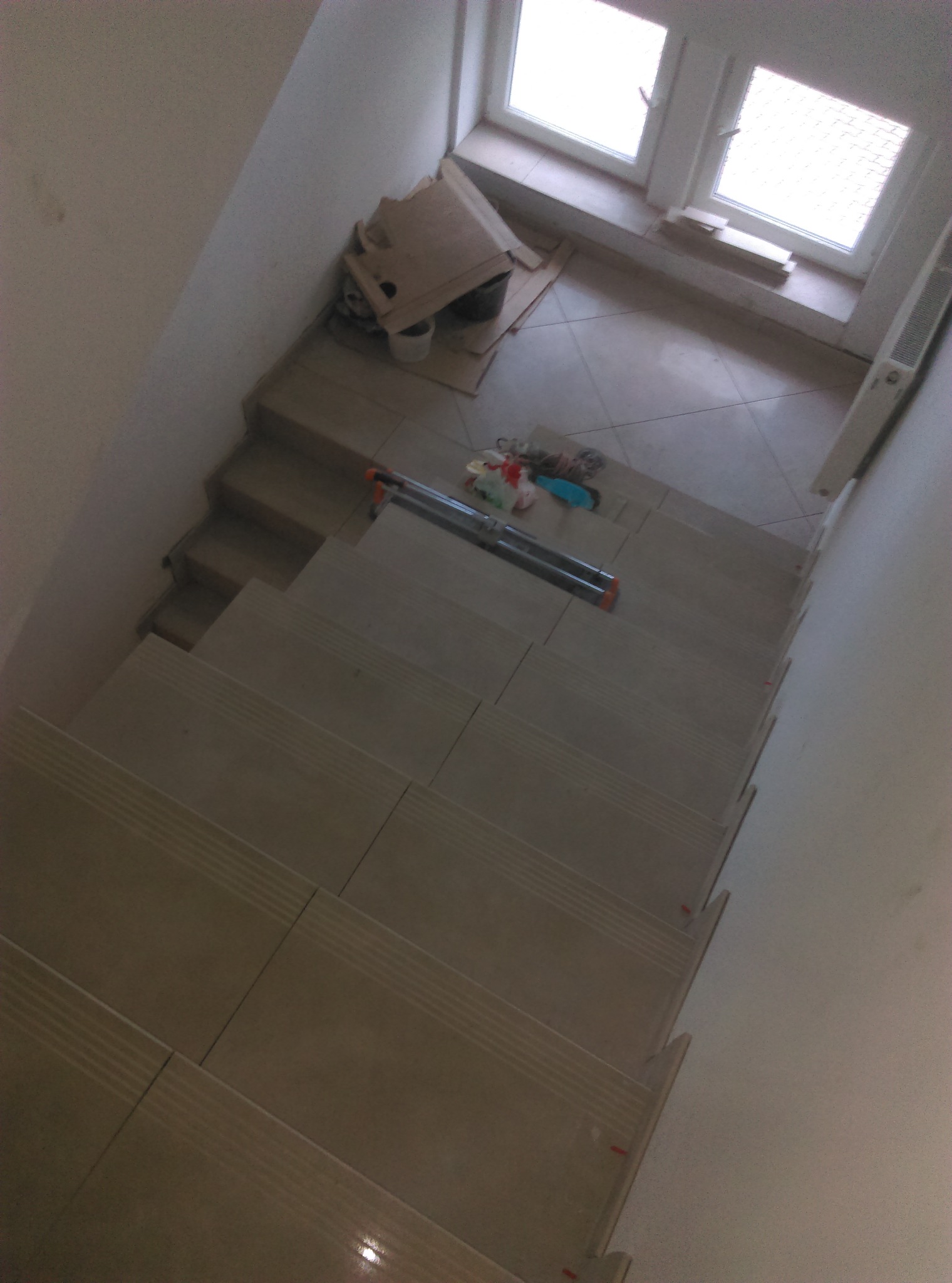 Klatka schodowa w trakcie wykańczania z ułożonymi płytkami na schodach, widoczne narzędzia i materiały budowlane, okno w tle