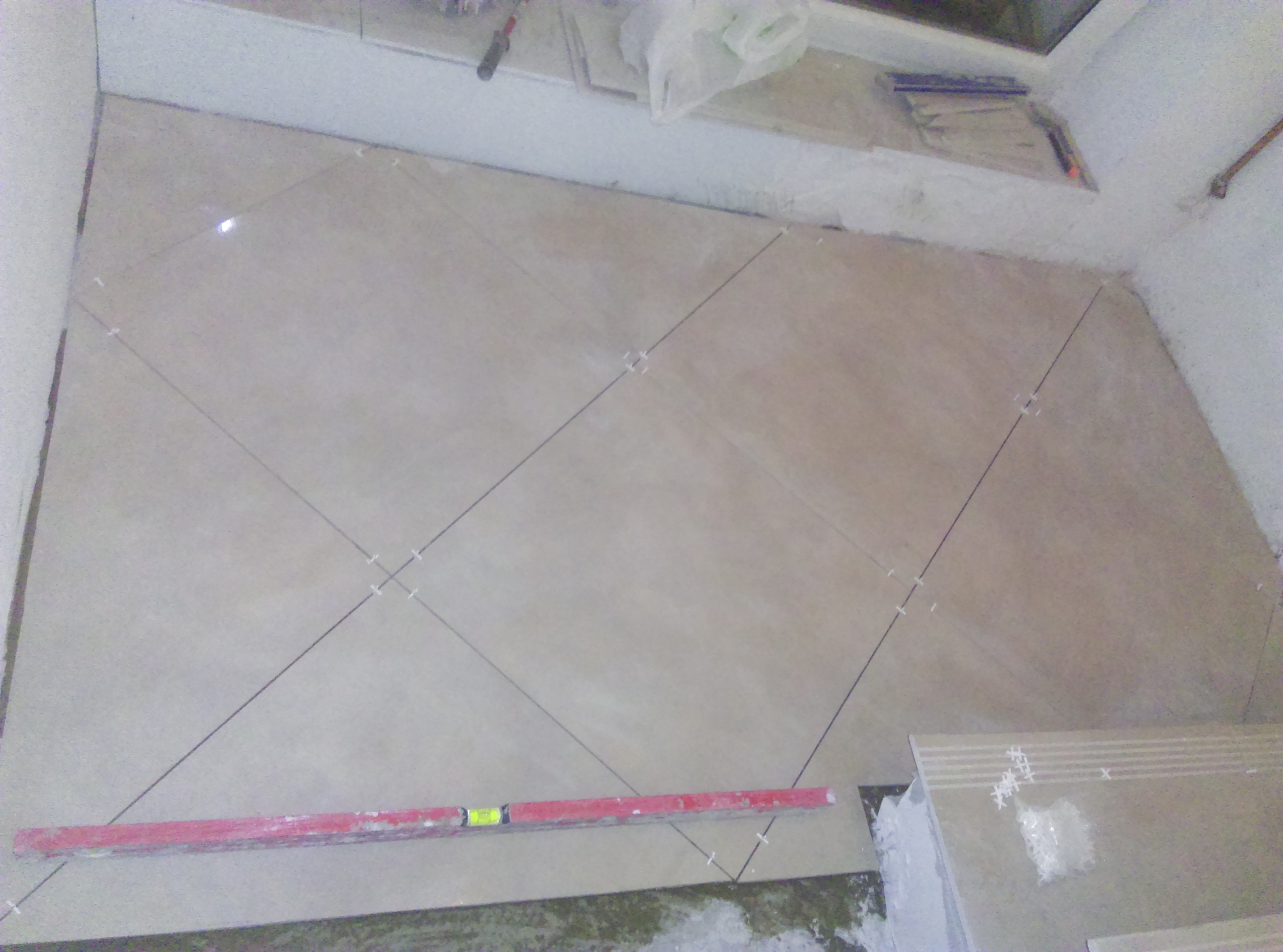 Ukośne układanie beżowych płytek podłogowych z użyciem krzyżyków dystansowych, widoczna poziomica i zapasowe płytki.