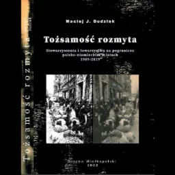 Okładka książki 'Tożsamość rozmyta' Macieja J. Dudziaka, przedstawiająca czarno-białe zdjęcie tłumu ludzi na tle budynków, z tekstem o stowarzyszeniach polsko-niemieckich w latach 1989-2019, wydana...