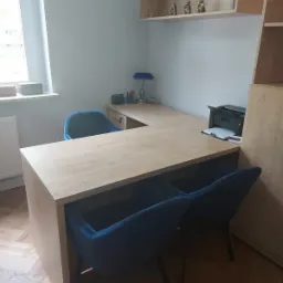 Gabinet z biurkiem w kształcie litery L, dwoma niebieskimi krzesłami, lampką na biurku i figurkami dekoracyjnymi na półce.