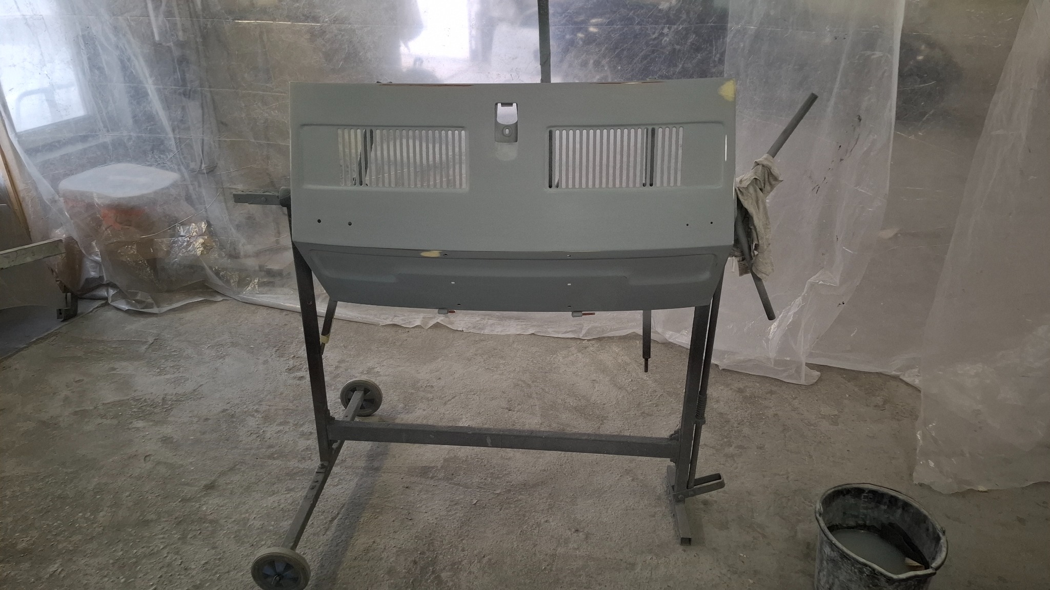 Przygotowana do lakierowania karoseria samochodu na metalowym stojaku w warsztacie. Widoczne ślady szpachli i przygotowania podkładu. Tło stanowi folia ochronna.