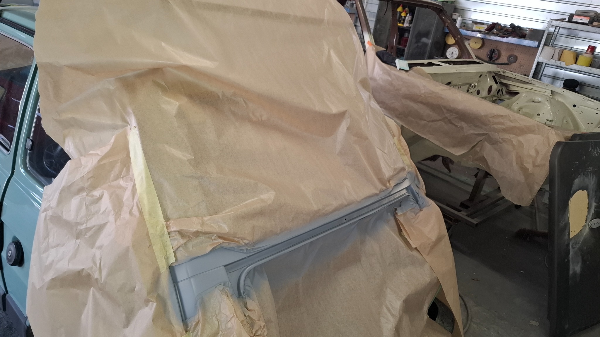 Przygotowanie do lakierowania: fragment karoserii samochodu, zabezpieczony papierem maskującym, świeżo pomalowany na szaro, widoczne elementy wnętrza i inne części pojazdu w tle.