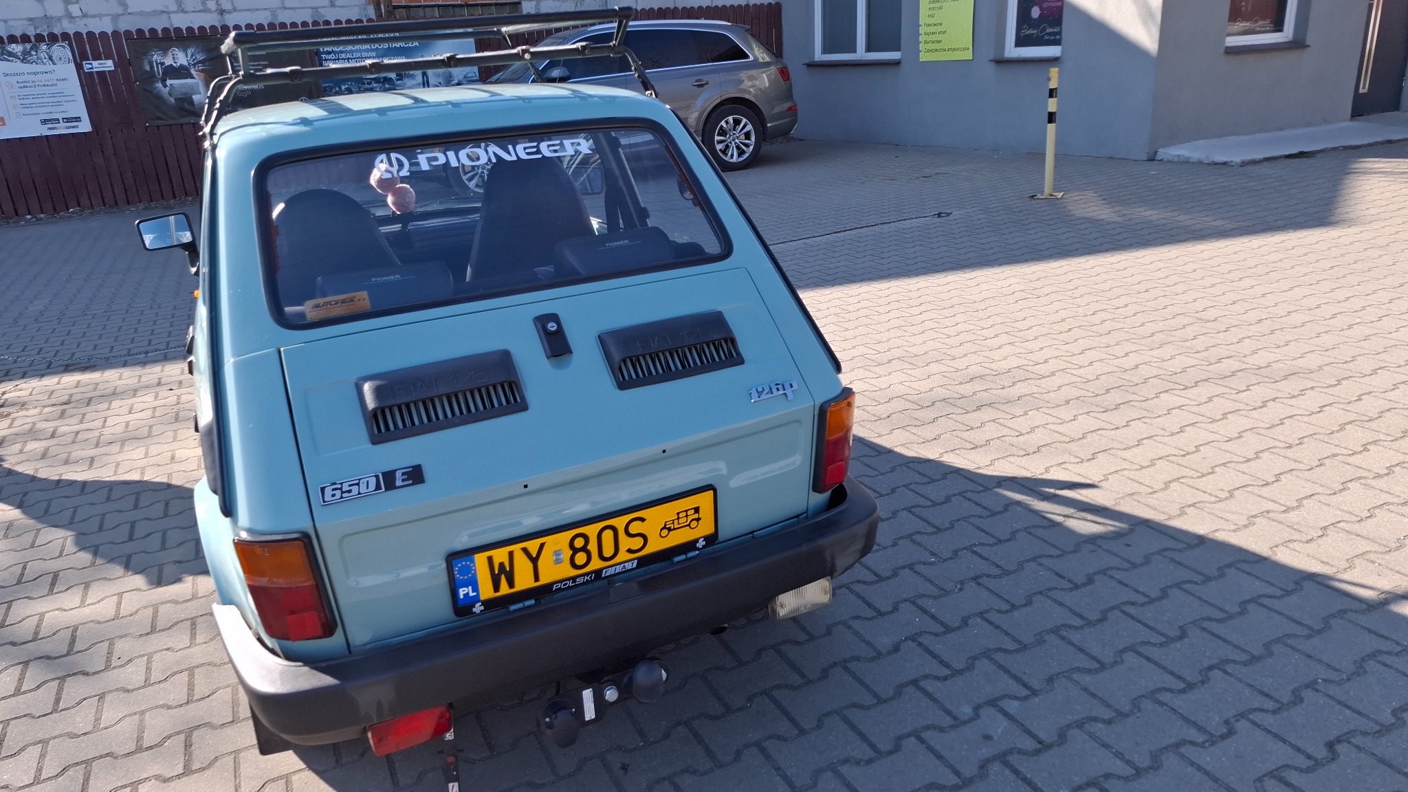 Błękitny Fiat 126p z bagażnikiem dachowym i naklejką 'Pioneer' na tylnej szybie, zaparkowany na brukowanej nawierzchni, z widocznym fragmentem innego auta w tle.