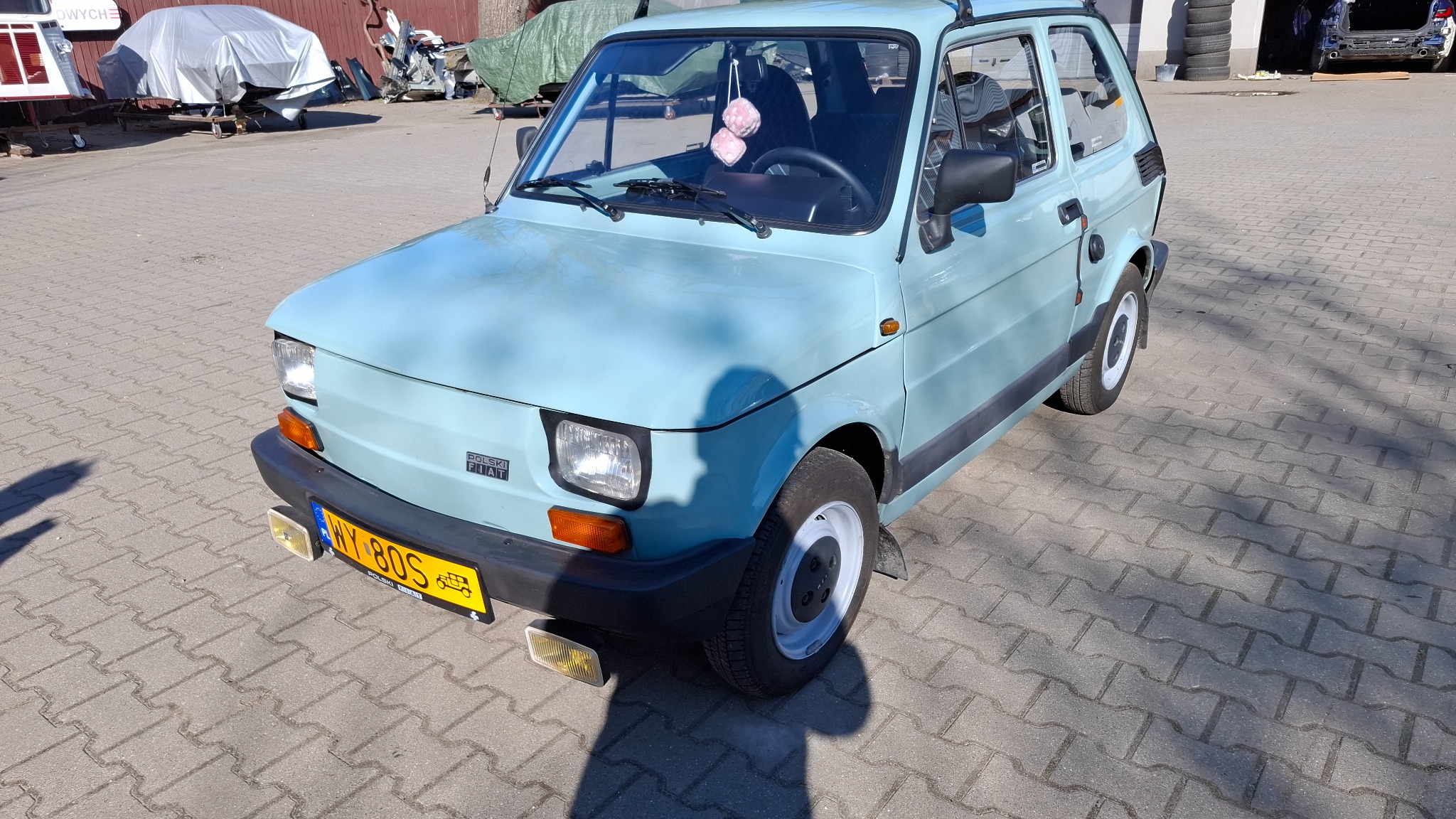 Jasnoniebieski Fiat 126p po renowacji, z białymi felgami i pluszowymi kostkami w środku, zaparkowany na brukowanym placu. Widoczne detale: tablica rejestracyjna.