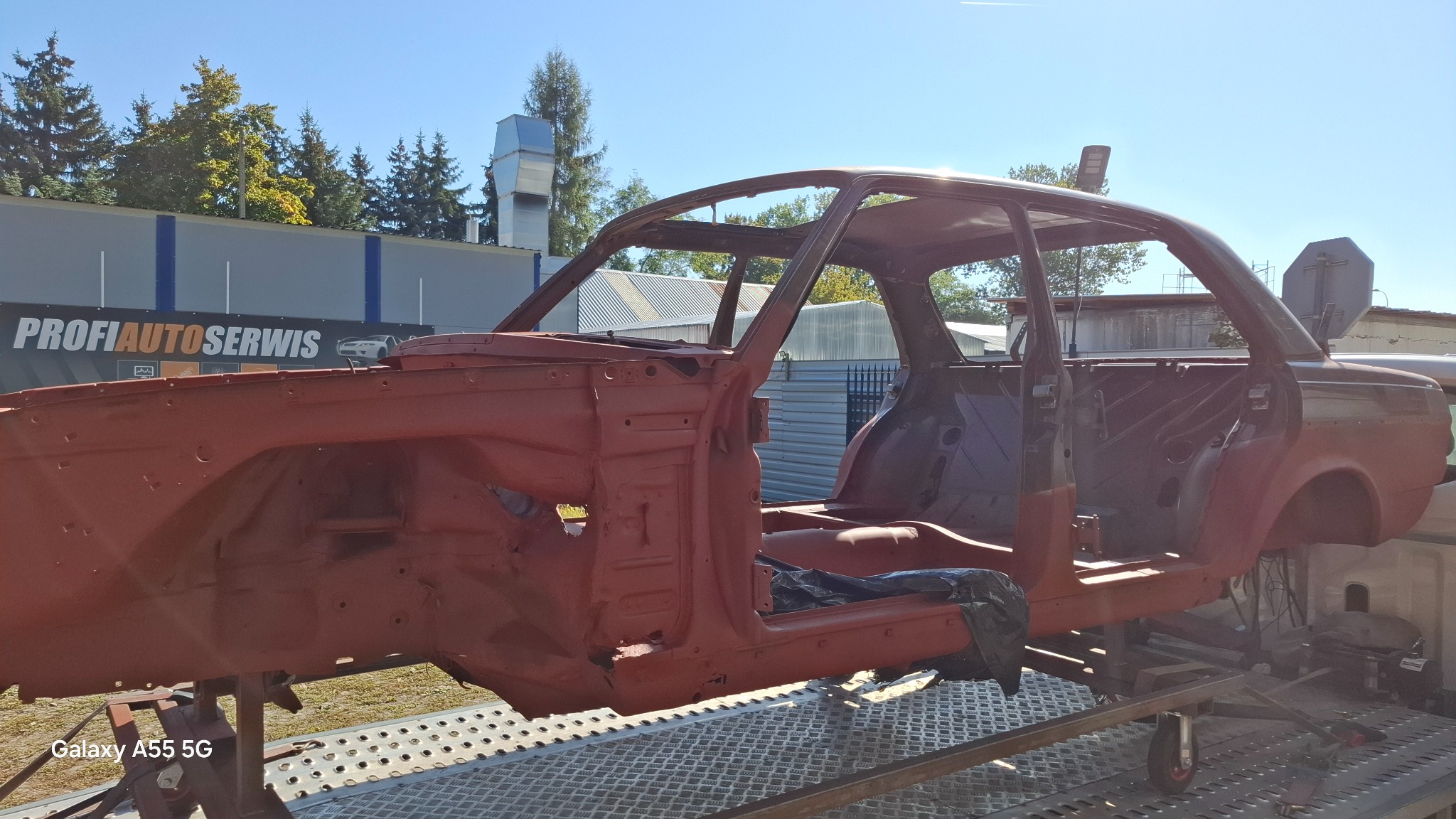 Czerwony szkielet karoserii samochodu na metalowej platformie, widok z boku, w tle budynek warsztatu Profi Auto Serwis, niebo i drzewa. Karoseria przygotowana do dalszych prac...