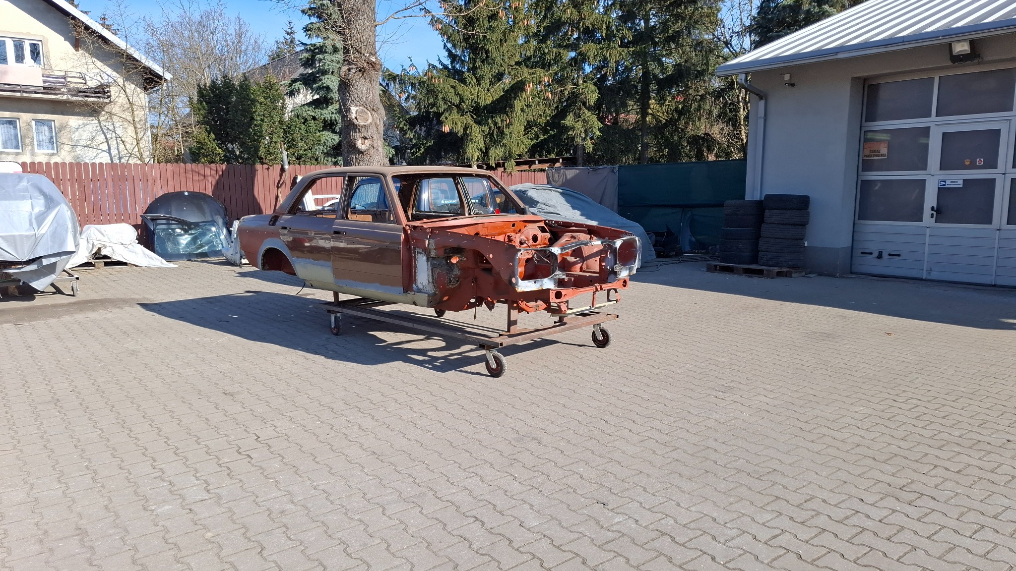 Karoseria samochodu na wózku w warsztacie w Warszawie, przygotowana do prac blacharsko-lakierniczych. Widoczne elementy nadwozia i opony. Słoneczny dzień.