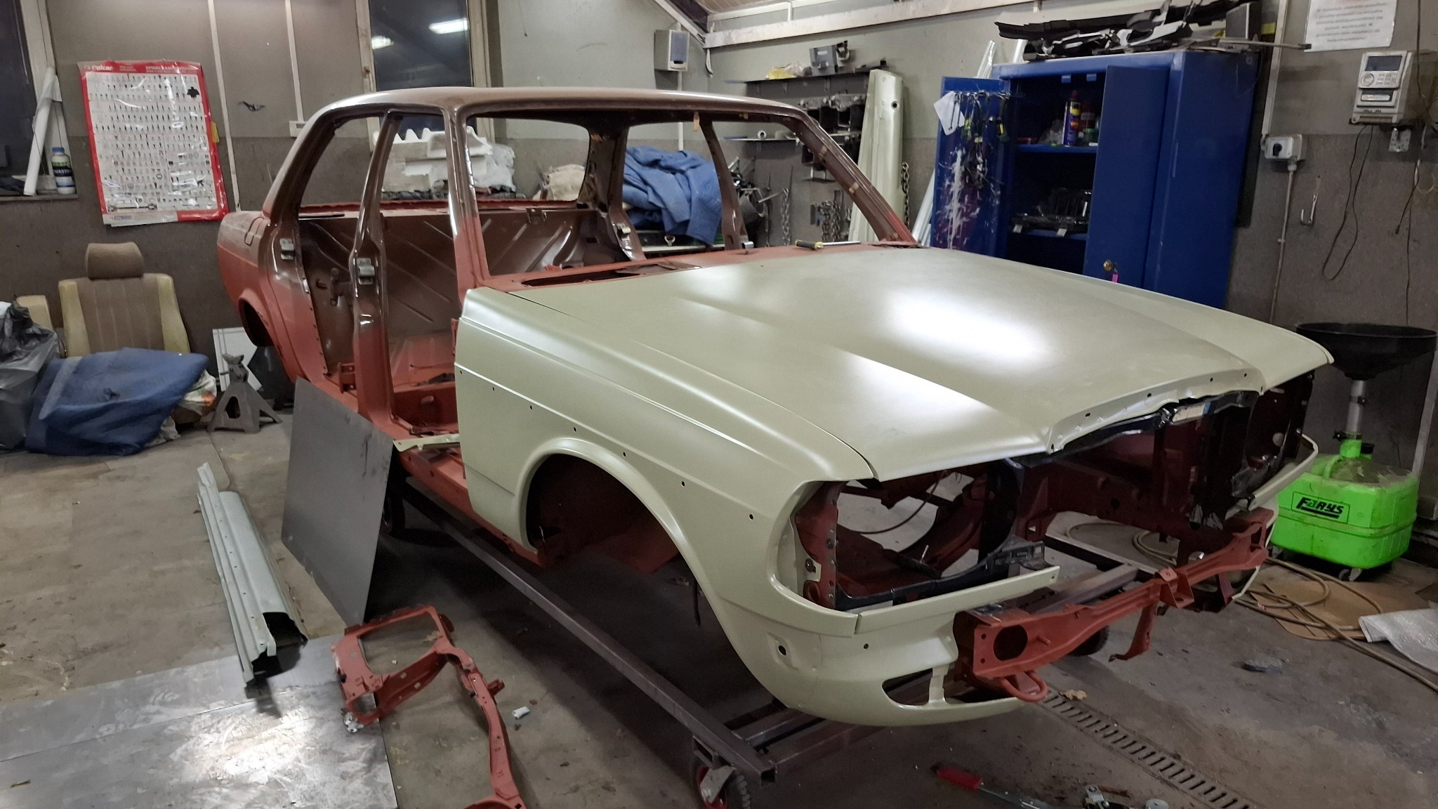 Karoseria klasycznego auta w trakcie renowacji blacharskiej i lakierniczej. Widoczne elementy w podkładzie i surowej blasze. Warsztat samochodowy w tle.