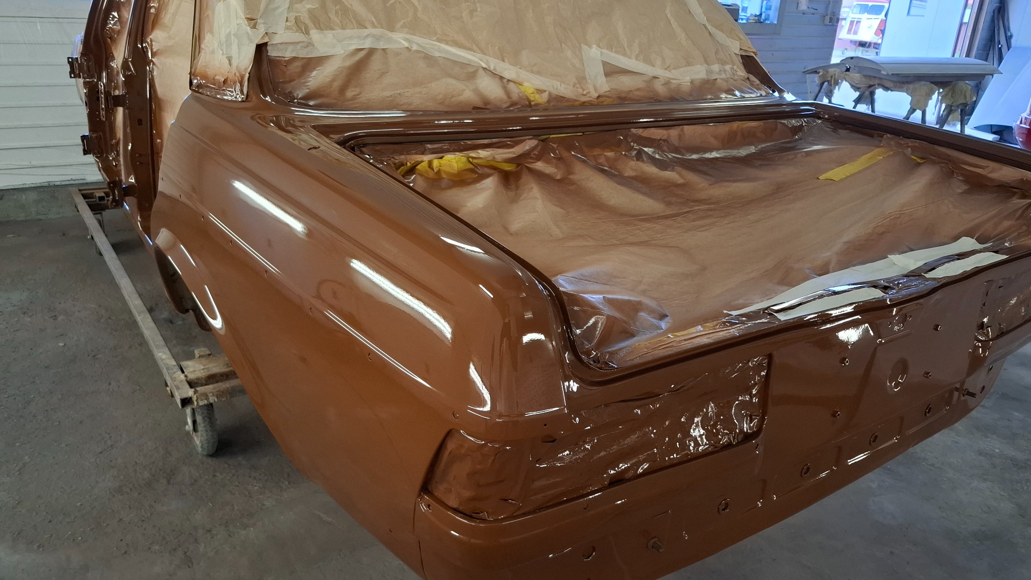 Błyszczący, brązowy lakier na karoserii samochodu w trakcie renowacji. Częściowo zamaskowany papierem i folią. Widoczne detale nadwozia i świeżo nałożona warstwa lakieru.