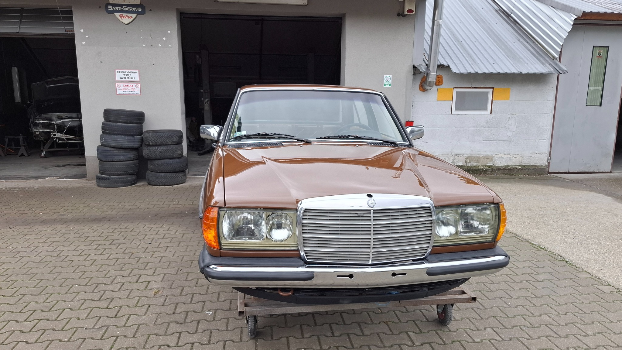 Brązowy Mercedes W123 na wózku warsztatowym przed budynkiem warsztatu. W tle sterta opon i częściowo widoczny szkielet innego pojazdu wewnątrz warsztatu.