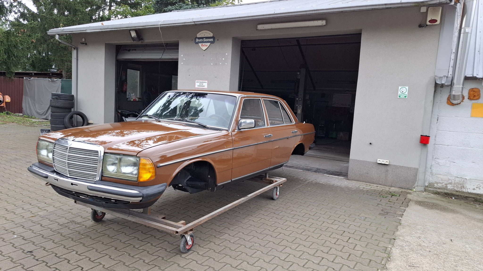 Brązowy Mercedes W123 bez kół, na wózku warsztatowym przed garażem z otwartymi wrotami. Widoczny podnośnik samochodowy w tle. Auto w trakcie renowacji.