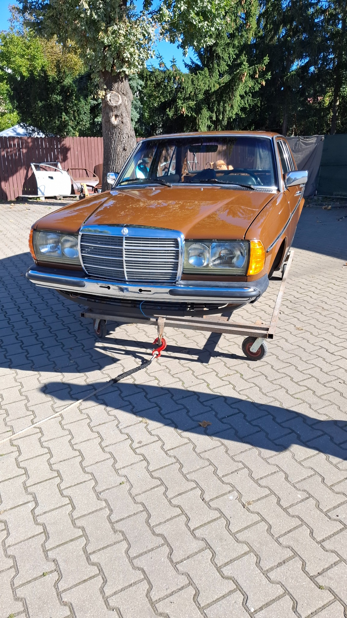 Brązowy Mercedes W123 na wózku warsztatowym, widok z przodu. W tle drzewo i fragment ogrodzenia. Auto przygotowane do prac blacharsko-lakierniczych.